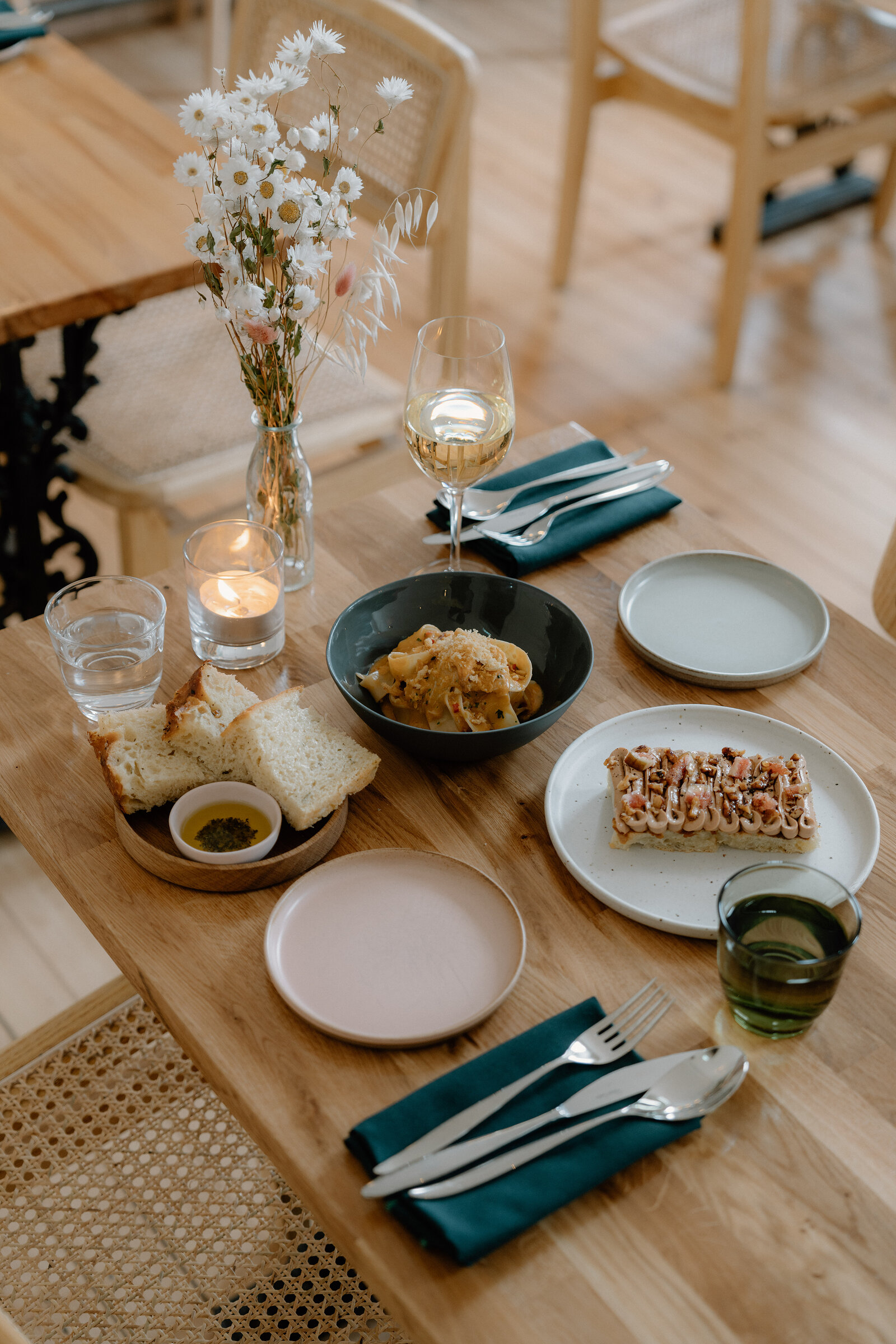 A restaurant table is set with a bowl of pasta, a plate of topped flatbread, a glass of white wine, sliced bread with olive oil, and a small vase of white daisies.