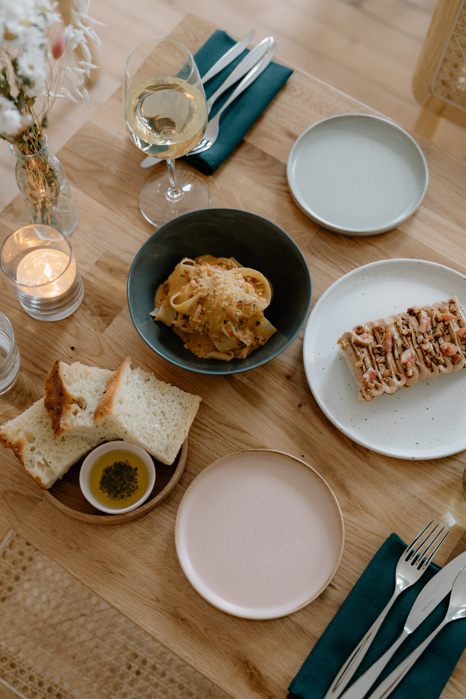 A wooden table holds a bowl of pasta with grated cheese, a plate of topped flatbread, a glass of white wine, and pale stoneware side plates with teal napkins.