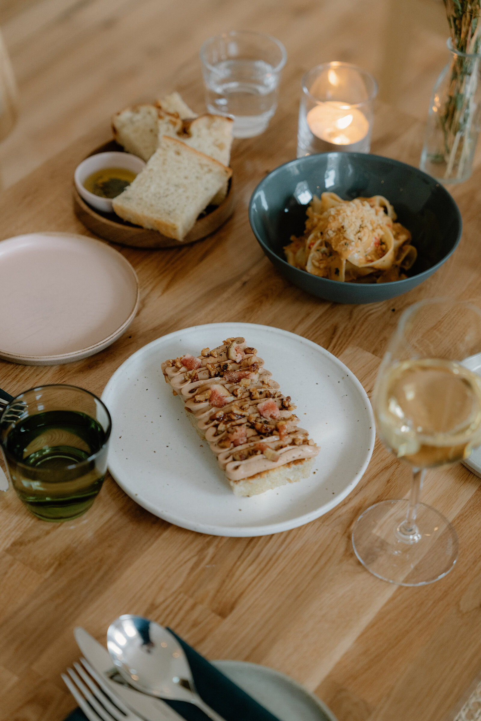 Sliced bread, a bowl of pasta, and a plate of topped flatbread sit on the wooden table at tipo, with a candle and a small vase of flowers behind.