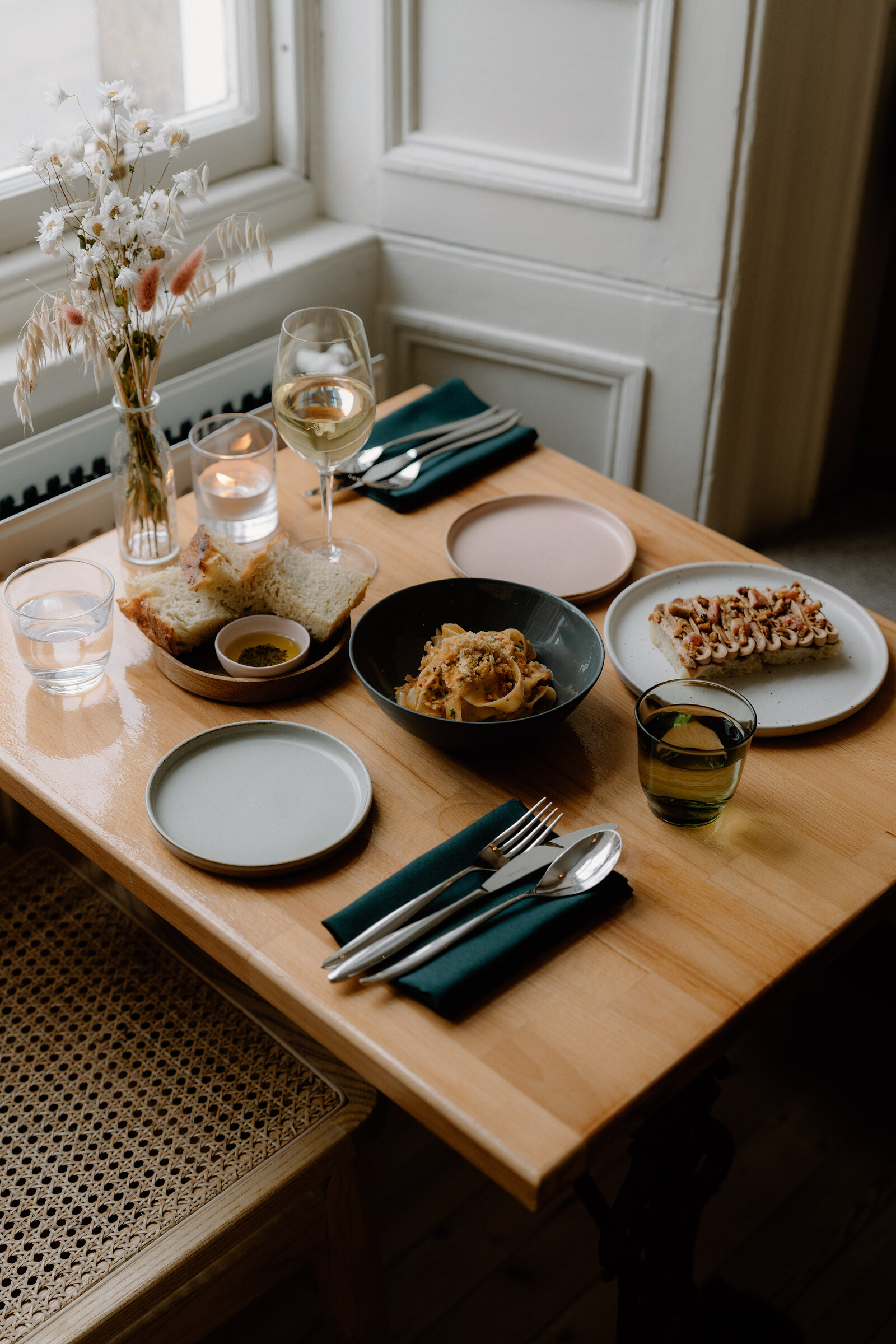 A table for two is laid by the window at tipo, with a bowl of pasta, a plate of topped flatbread, a glass of white wine, and a small vase of dried flowers.