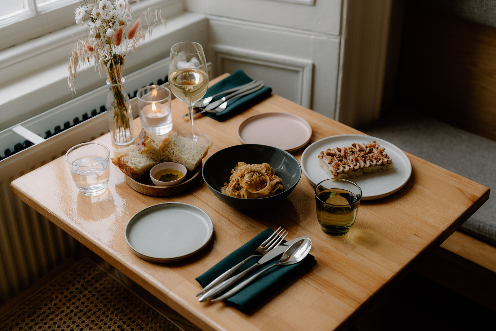 Plates of pasta and topped flatbread are set on a wooden table beneath a tall panelled window at tipo, with cane chairs and candlelight.