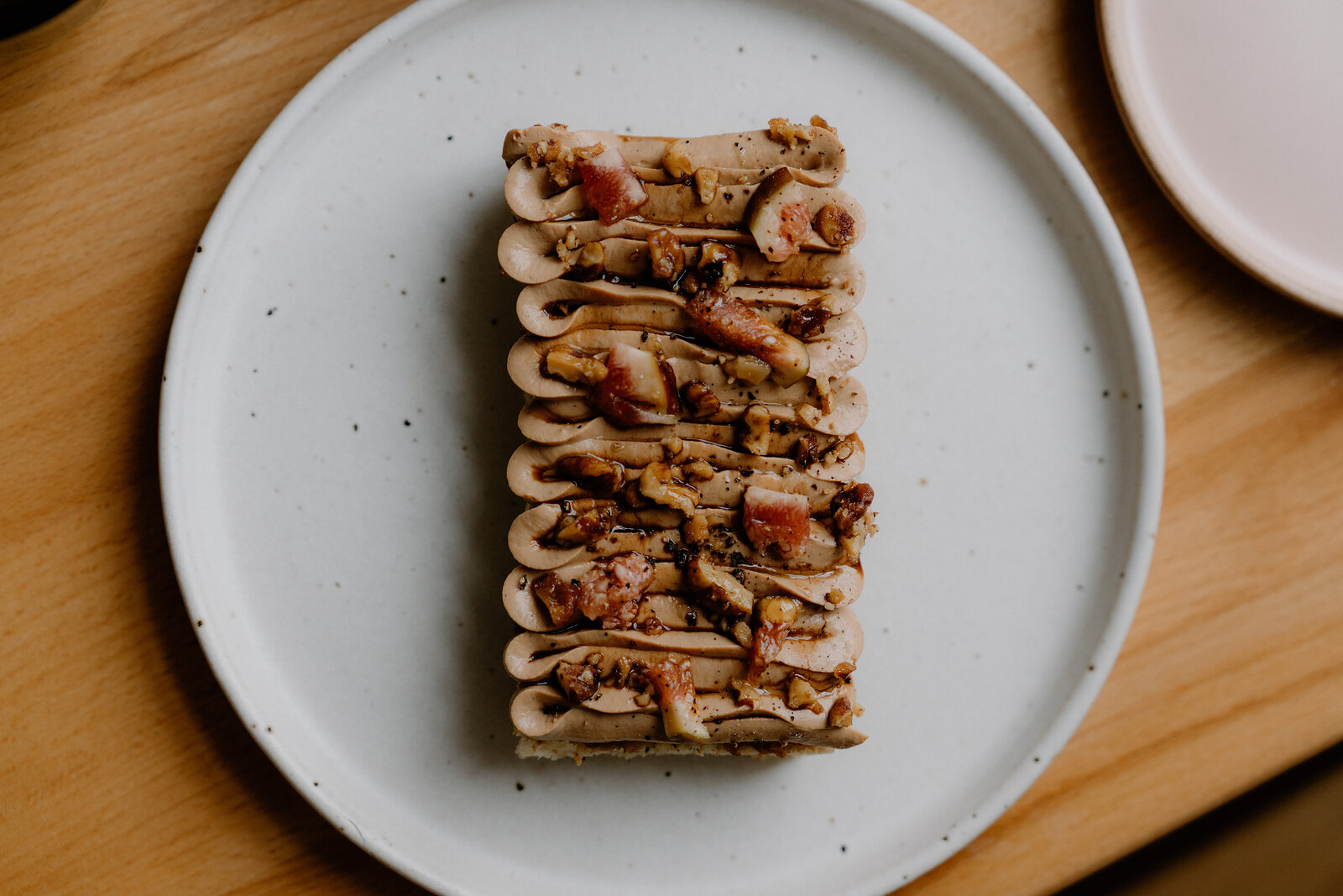 A rectangular parfait is piped with pale mousse in ridges, scattered with chopped nuts and small pink pieces, on a speckled white plate.
