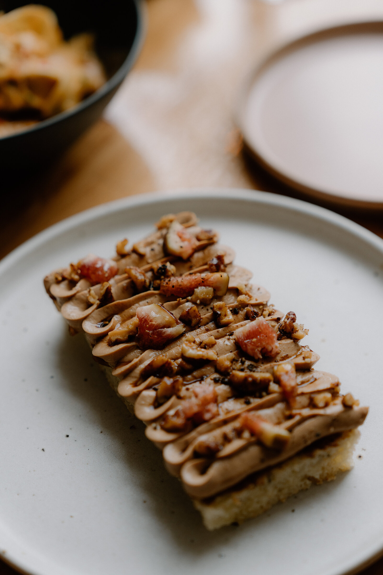 Chopped nuts and small pink pieces top a piped mousse terrine on a speckled plate, photographed close with a bowl of pasta blurred behind.