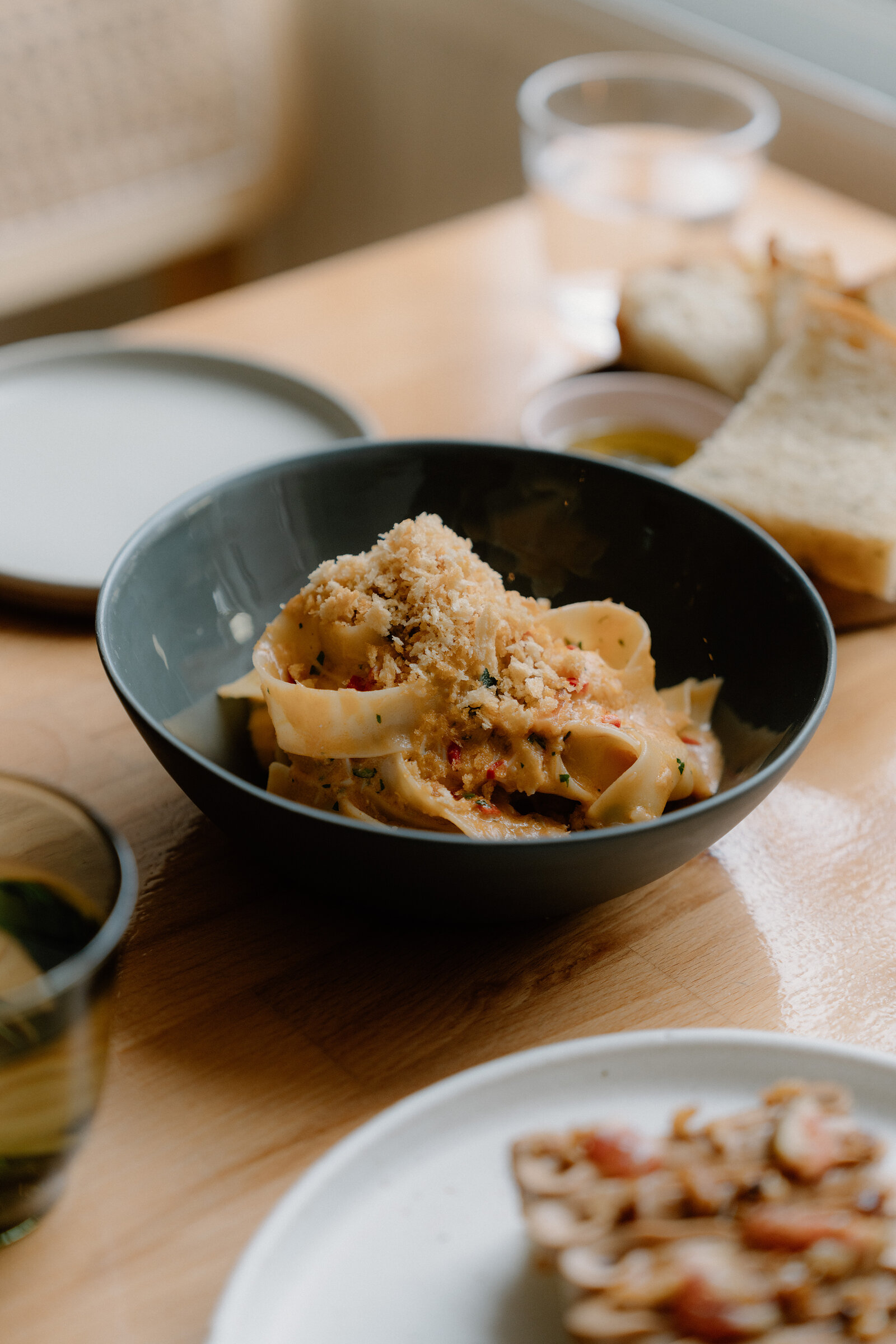 A dark bowl of tagliatelle topped with breadcrumbs sits on the wooden table at tipo, with sliced bread and a water glass behind.