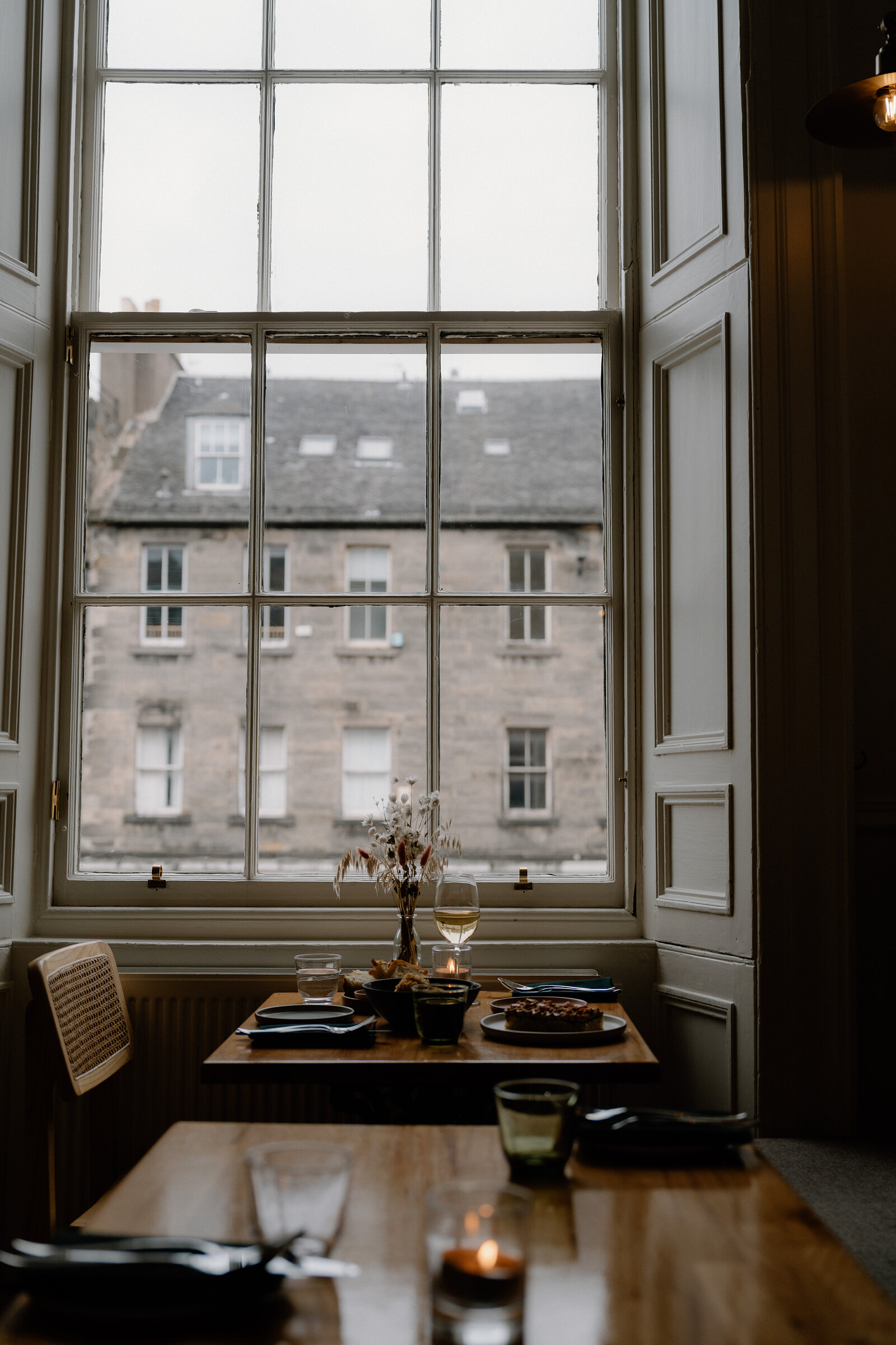 A window table at tipo looks out over Edinburgh tenement rooftops, laid with a candle, dried flowers, and glassware against wood panelling.
