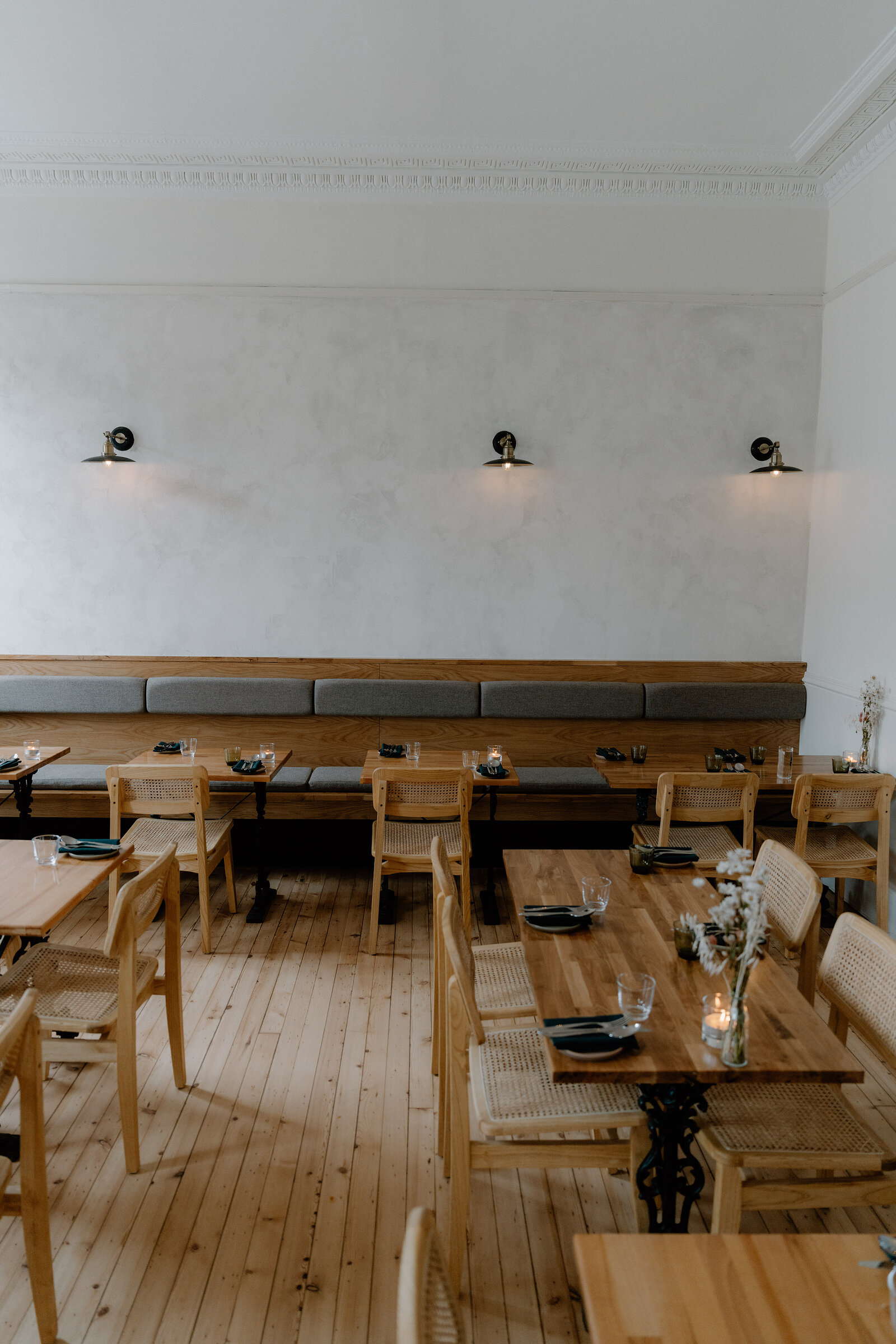 Wooden tables and chairs sit before a grey panelled wall lined with wall lamps and a long banquette seat in the tipo dining room.