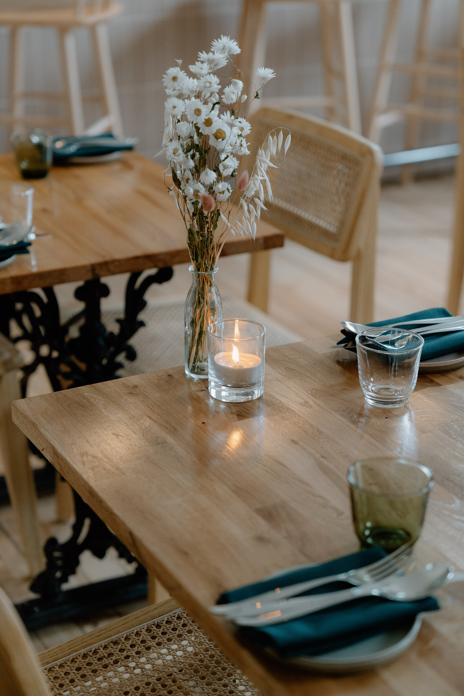 A small vase of white daisies and dried flowers stands beside a lit tealight candle on a wooden table at tipo, with green glassware and teal napkins set nearby.