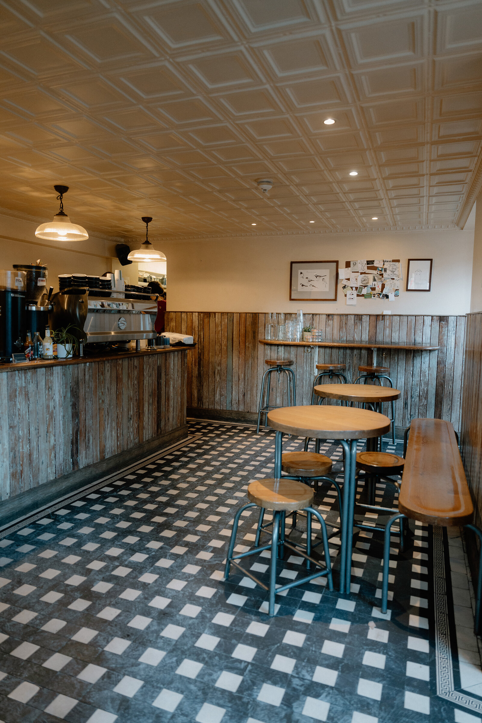 A patterned tin ceiling arches over an empty cafe seating area of round tables, stools and a wooden bench on a checkerboard floor.