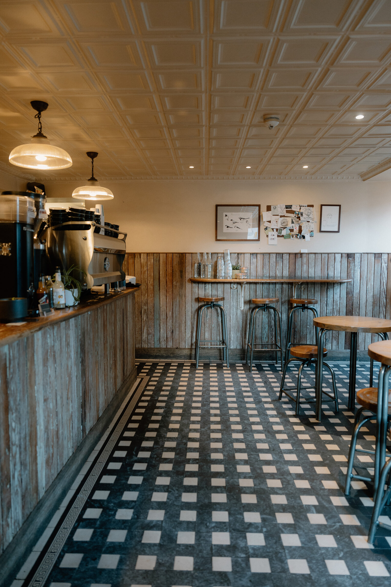 An espresso machine sits on a weathered wooden counter beneath a patterned tin ceiling, stools lined along the wall behind.