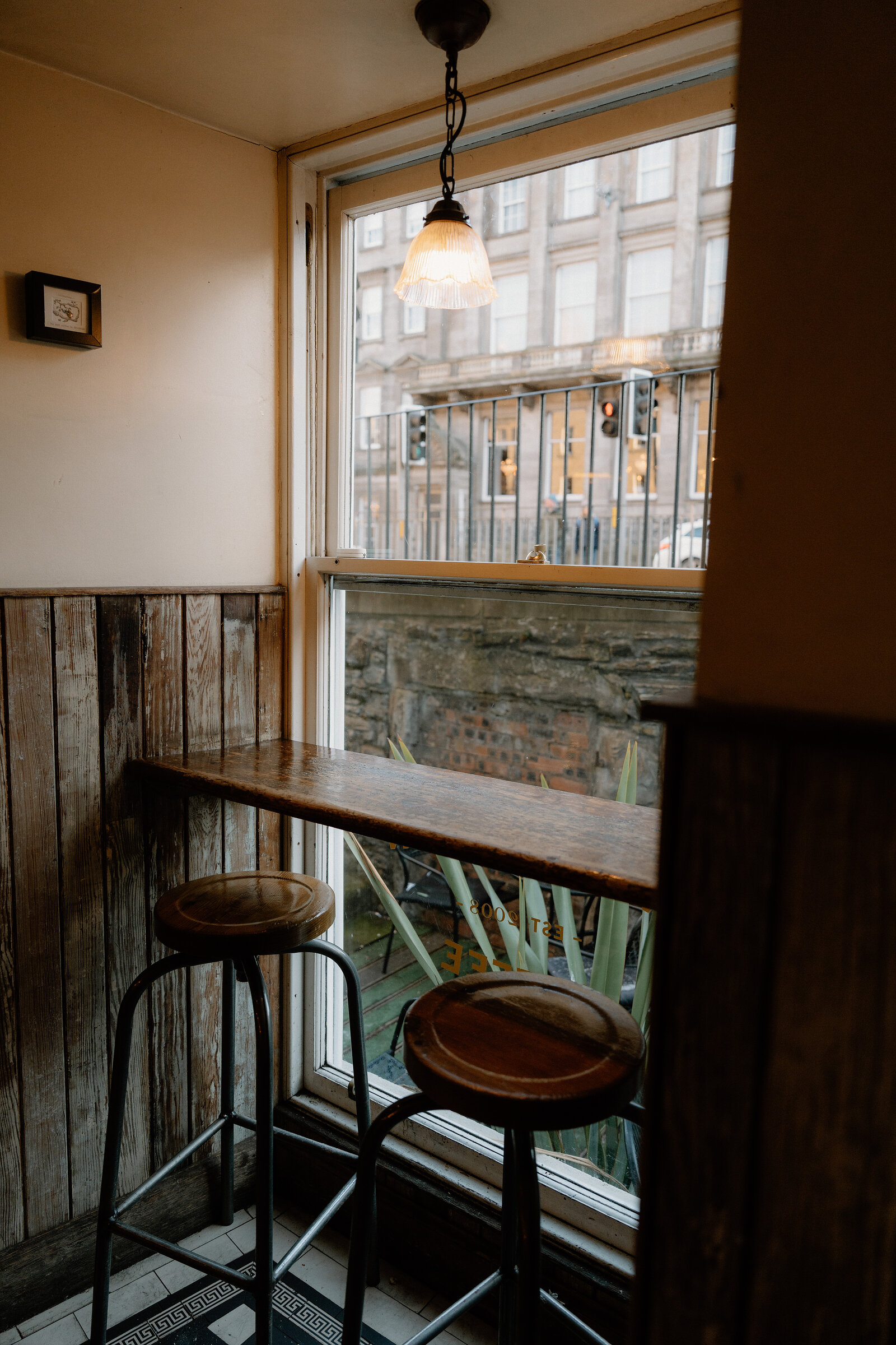 A wooden window bar with two stools looks out through etched glass onto a New Town street, a potted plant on the sill outside.