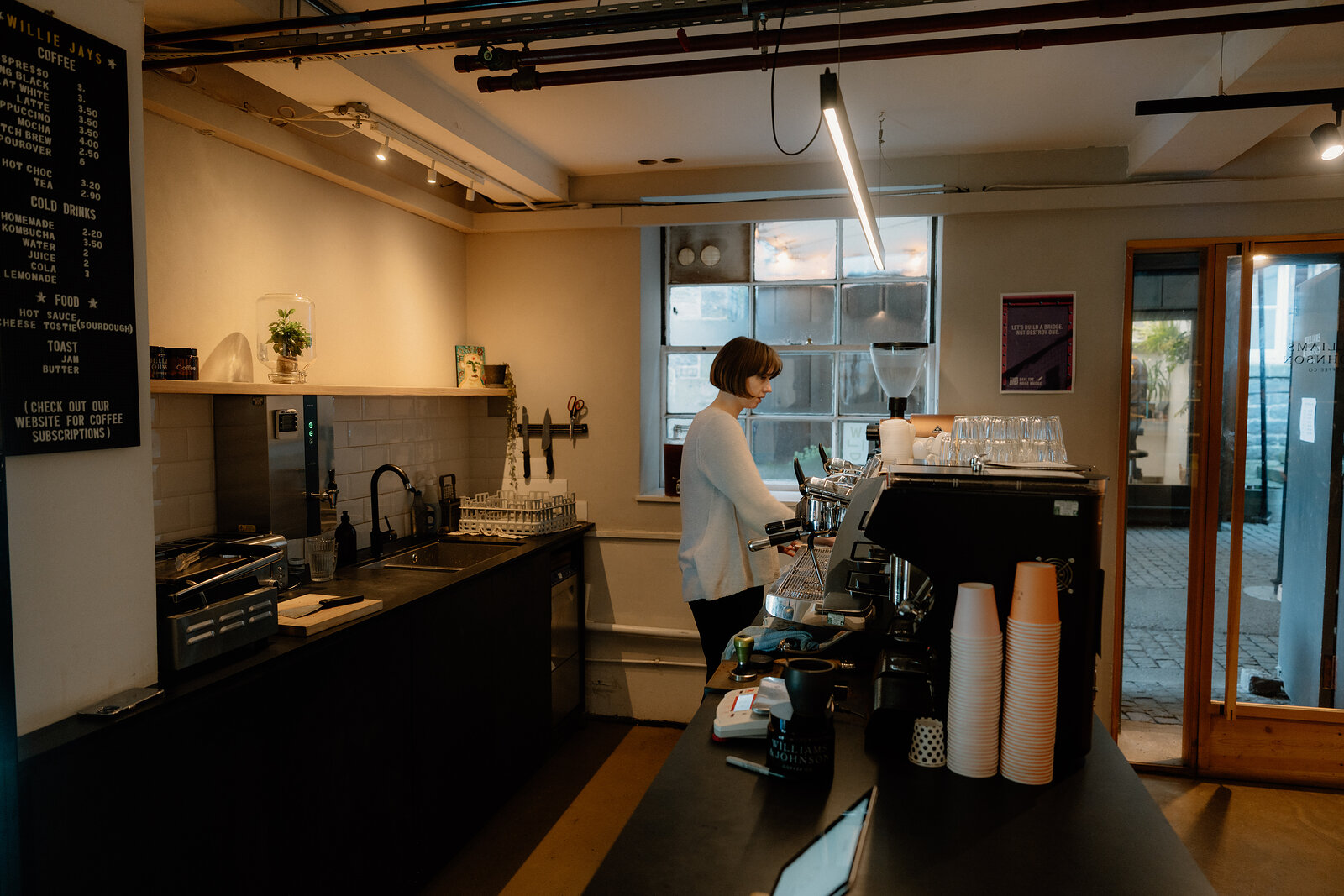Diners sit at small tables in a dimly lit cafe with exposed pipework, black pendant lamps and a large potted plant by the counter.