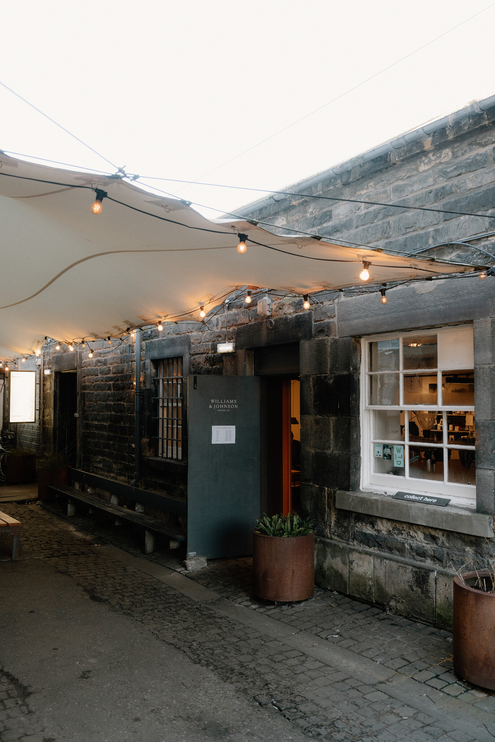 String lights hang beneath a canvas awning outside Williams & Johnson's dark stone building, potted plants flanking the entrance.