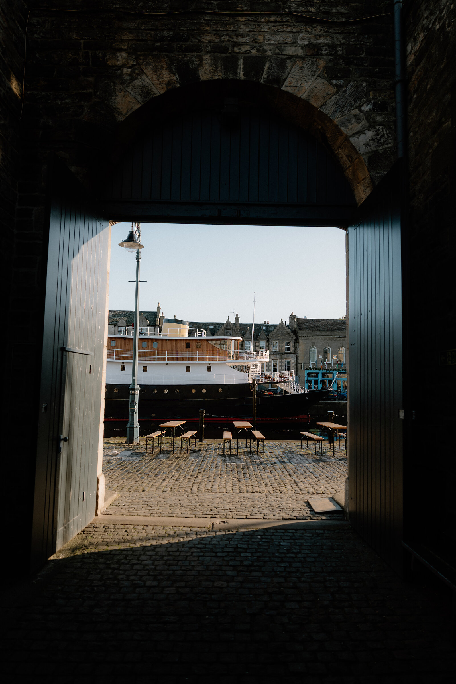 A dark stone archway frames a sunlit quayside where a moored ship and rows of benches sit on cobblestones.