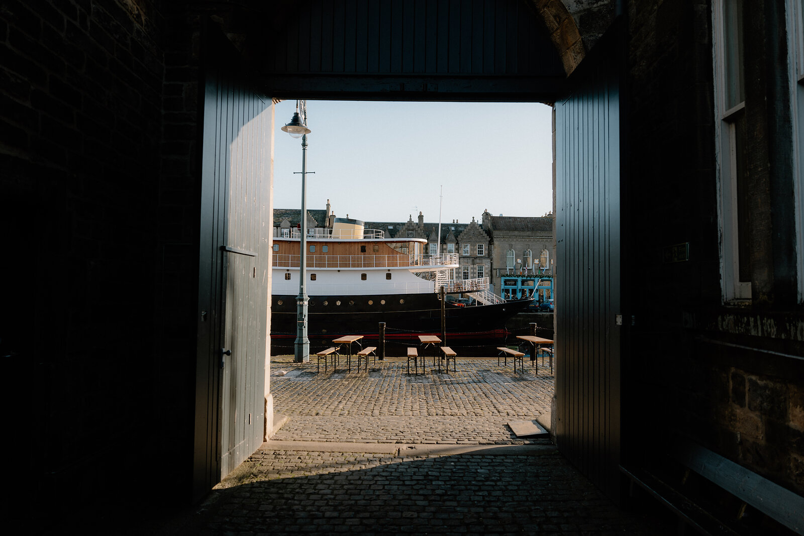 Sunlight floods through a warehouse archway onto a cobbled quay where a large ship is moored beside benches and a lamppost.