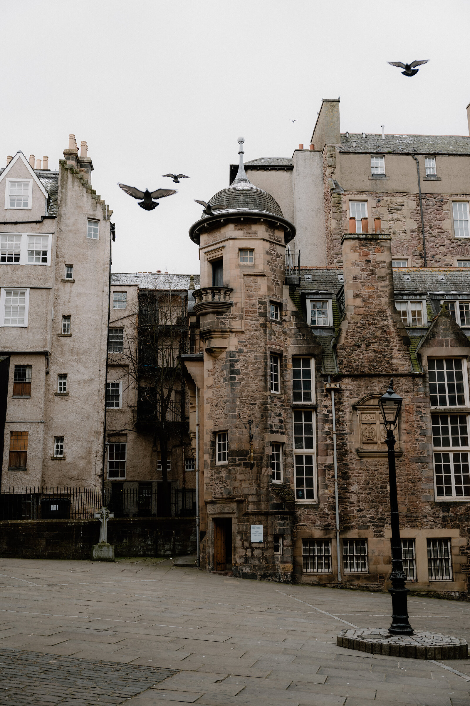 Pigeons scatter above a turreted sandstone building overlooking a quiet paved square lined with gabled tenements.
