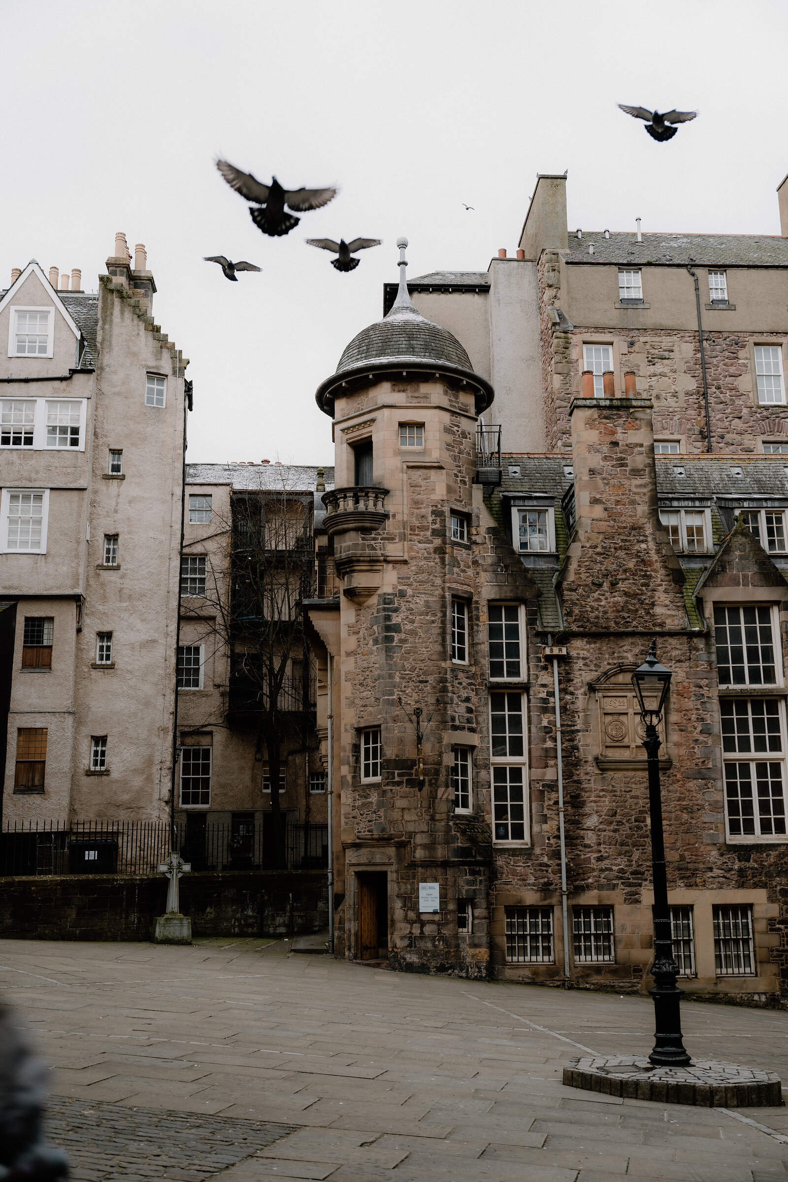 Birds fly above a conical-roofed stone turret set into a row of Gothic-windowed tenements around a paved square.
