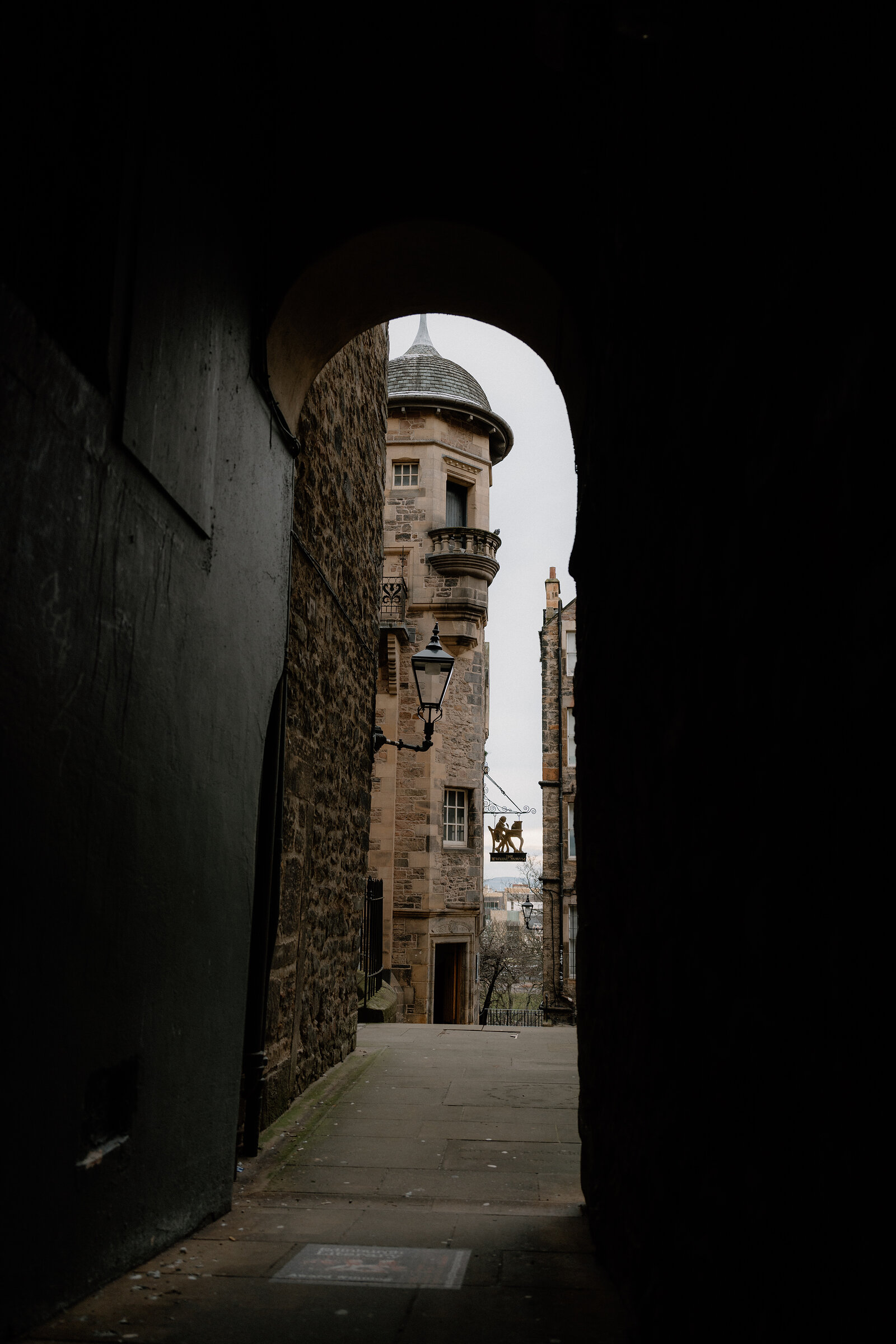 A stone archway frames a view down a narrow close toward a domed turret and a hanging pub sign.