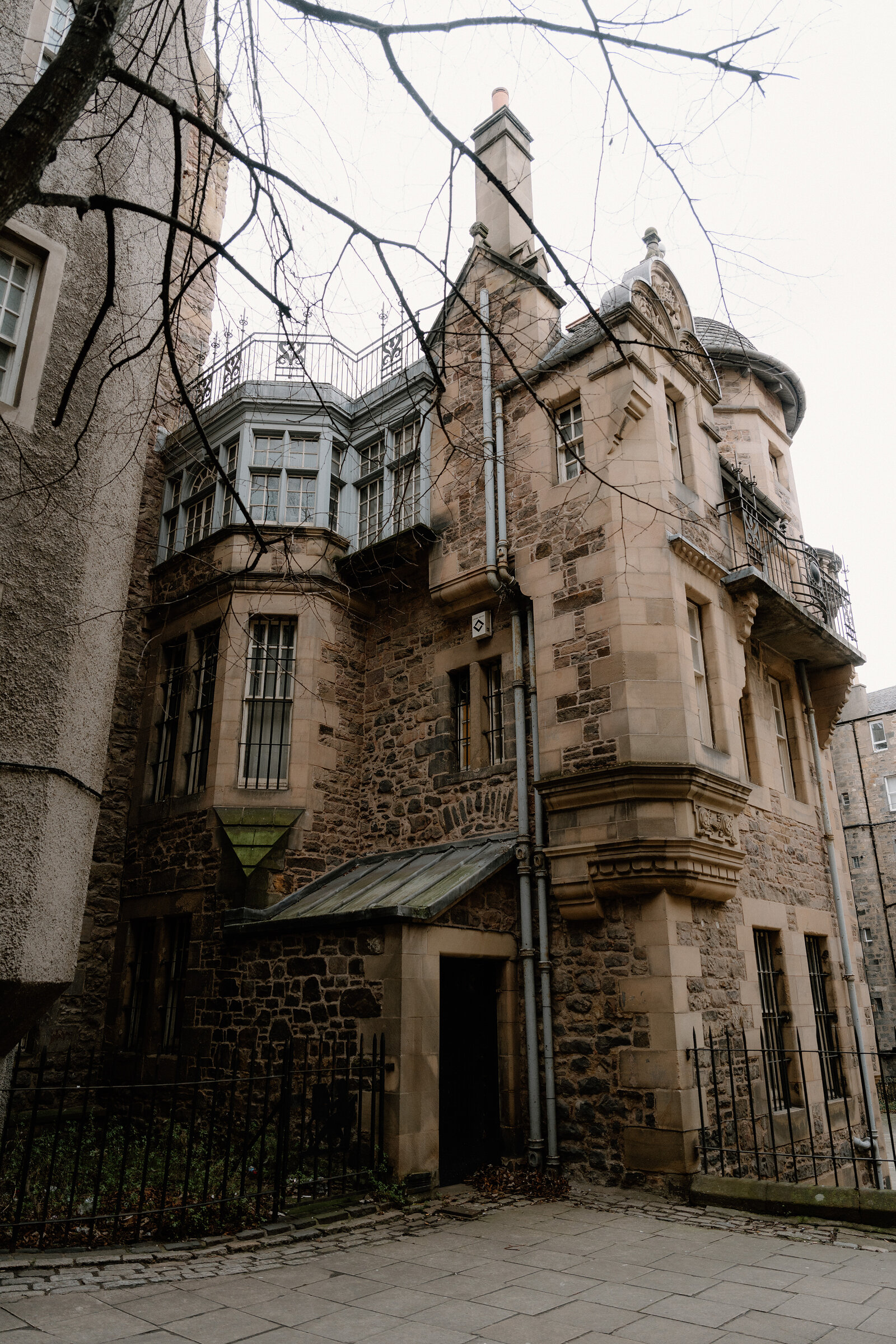 An ornate sandstone townhouse with bay windows, a round corner turret and a covered entrance porch stands beside bare winter trees.
