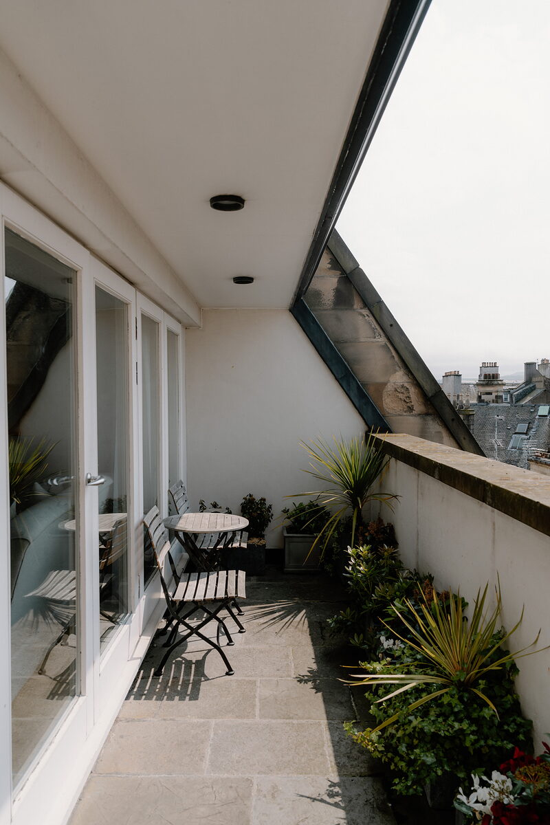 A rooftop terrace with a bistro table and chairs among potted plants, overlooking the city.