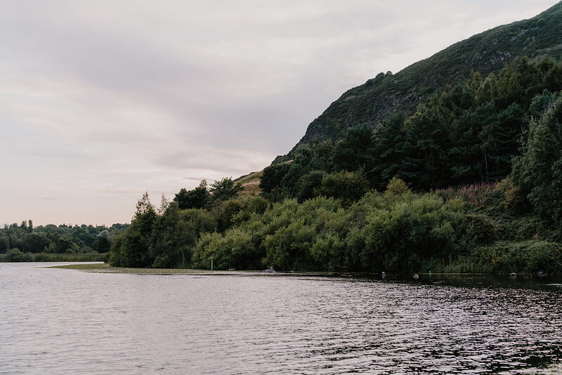 Duddingston Loch's shoreline curves beneath a wooded hillside, the water calm and still.