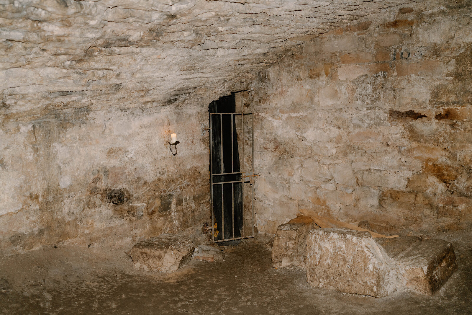 A barred iron gate set into a rough stone chamber underground, a lantern lighting the wall beside it