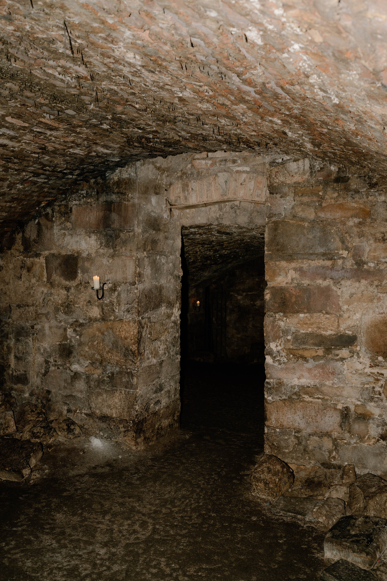 A vaulted stone passage opening into a darker chamber underground, a lantern lighting the wall