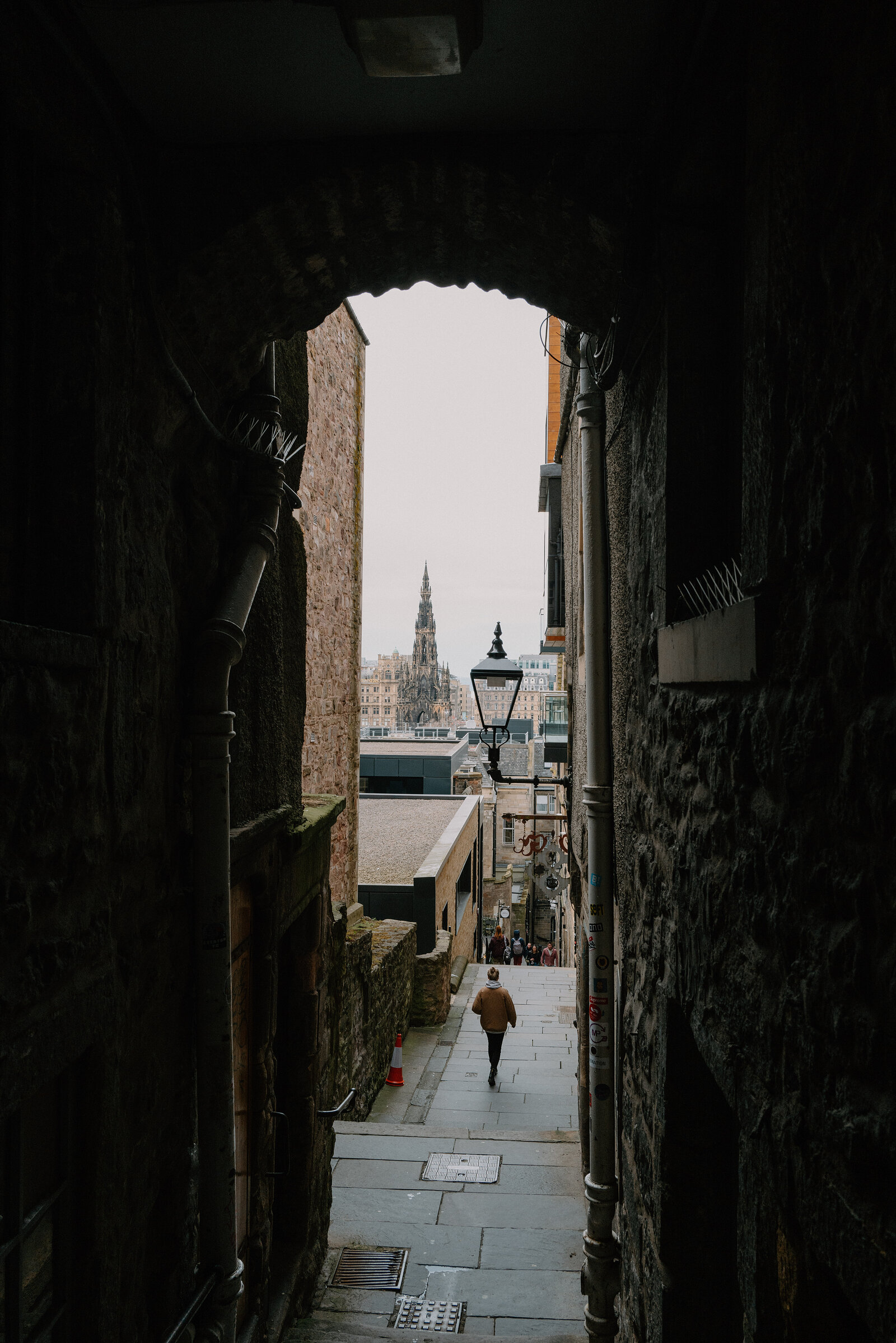 A pedestrian walking down a narrow Old Town close, the Scott Monument's spire rising beyond the rooftops.