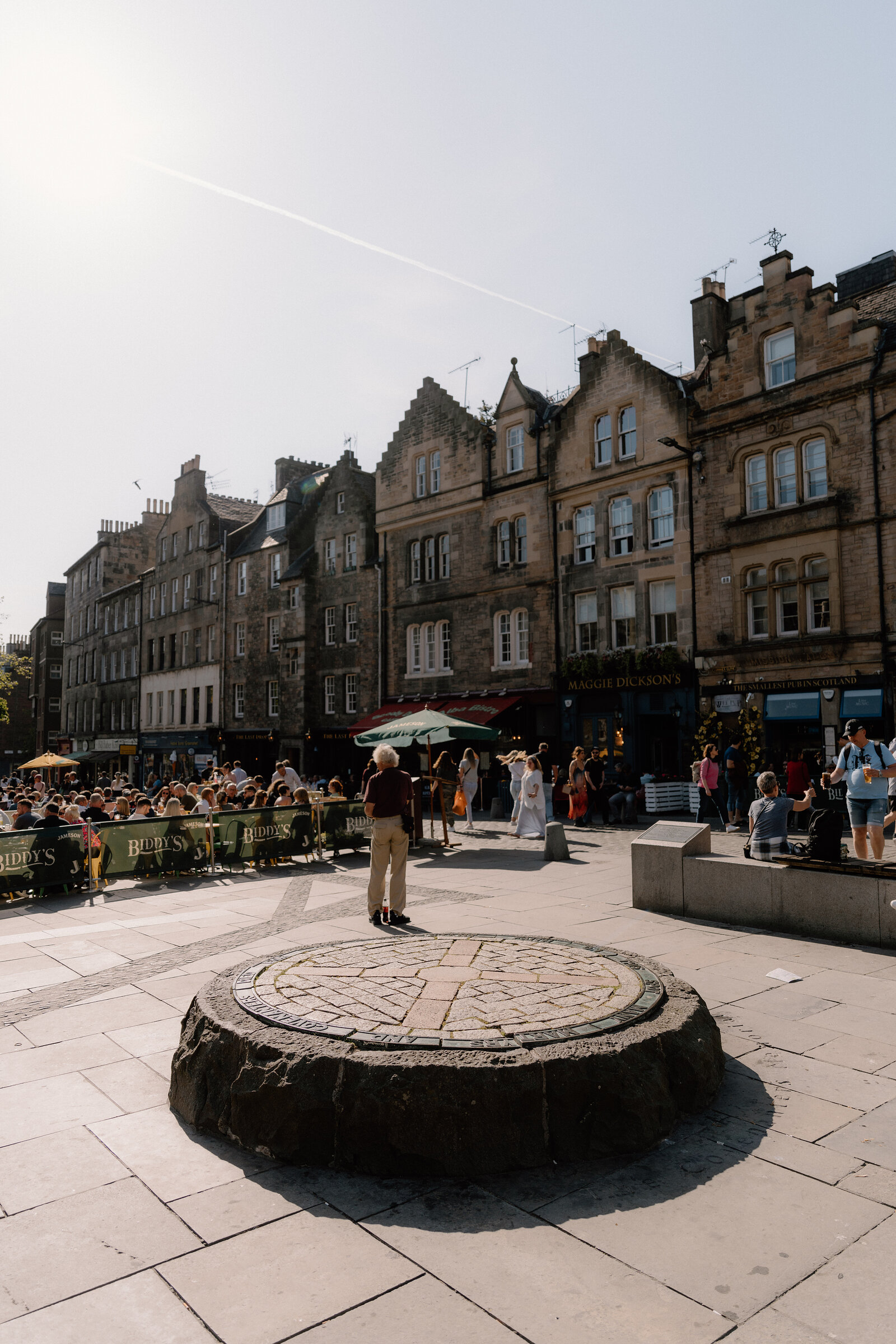 A cobbled square in Edinburgh's Old Town, crowds gathered near a circular stone marker beneath tall tenements.