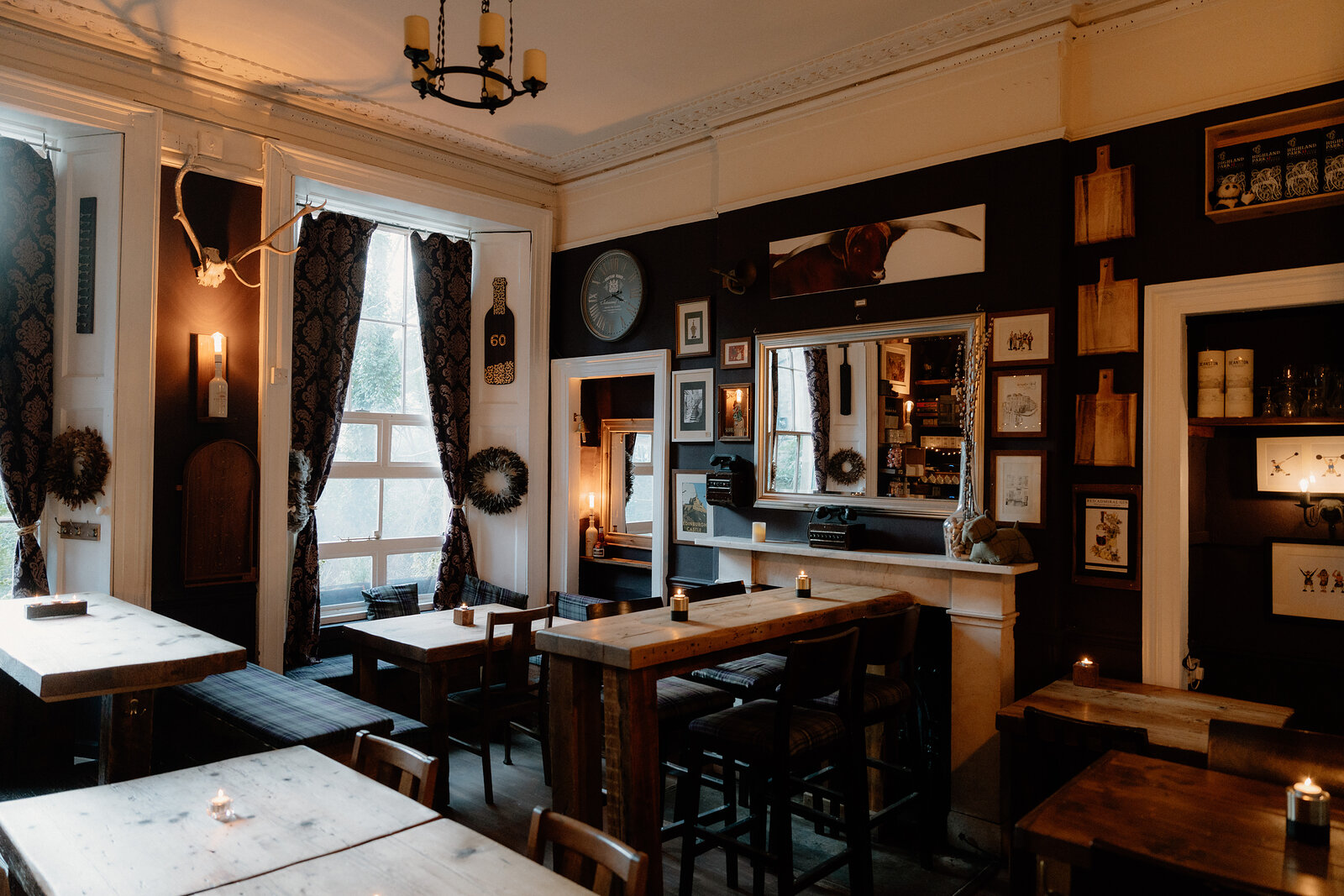 A dark dining room with mirrors and framed prints above a fireplace, wreaths hanging beside curtained windows