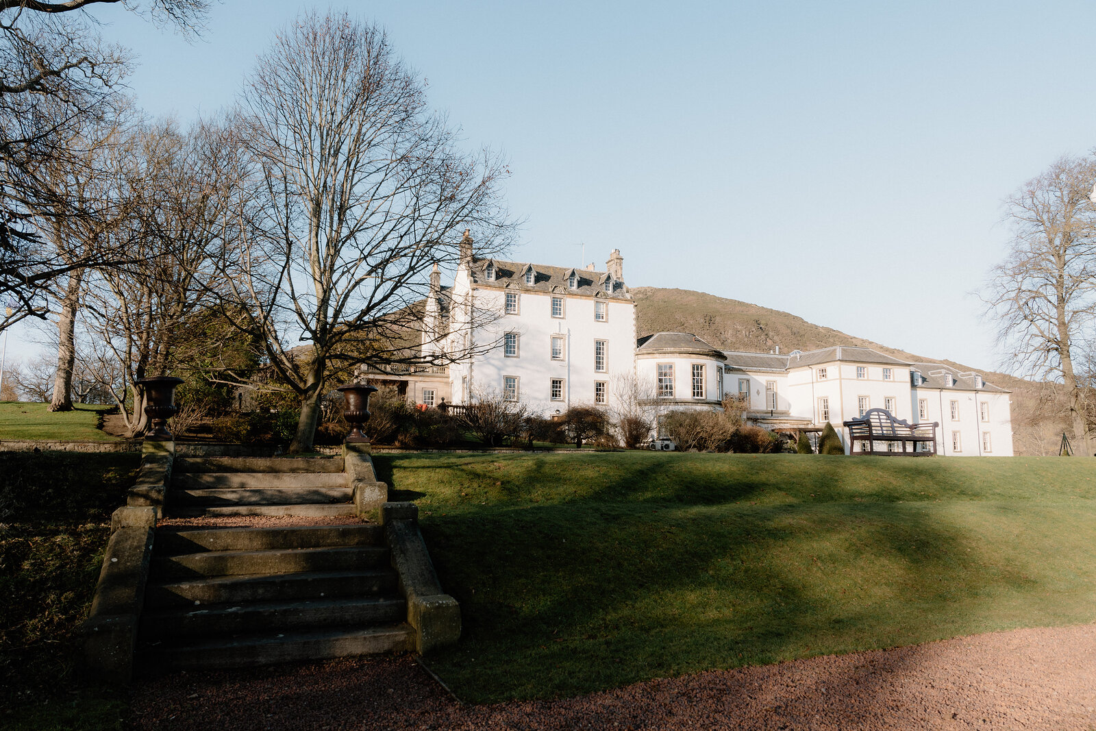 Prestonfield House's white facade rises behind stone steps and mature trees, Arthur's Seat's slopes visible on the skyline beyond.