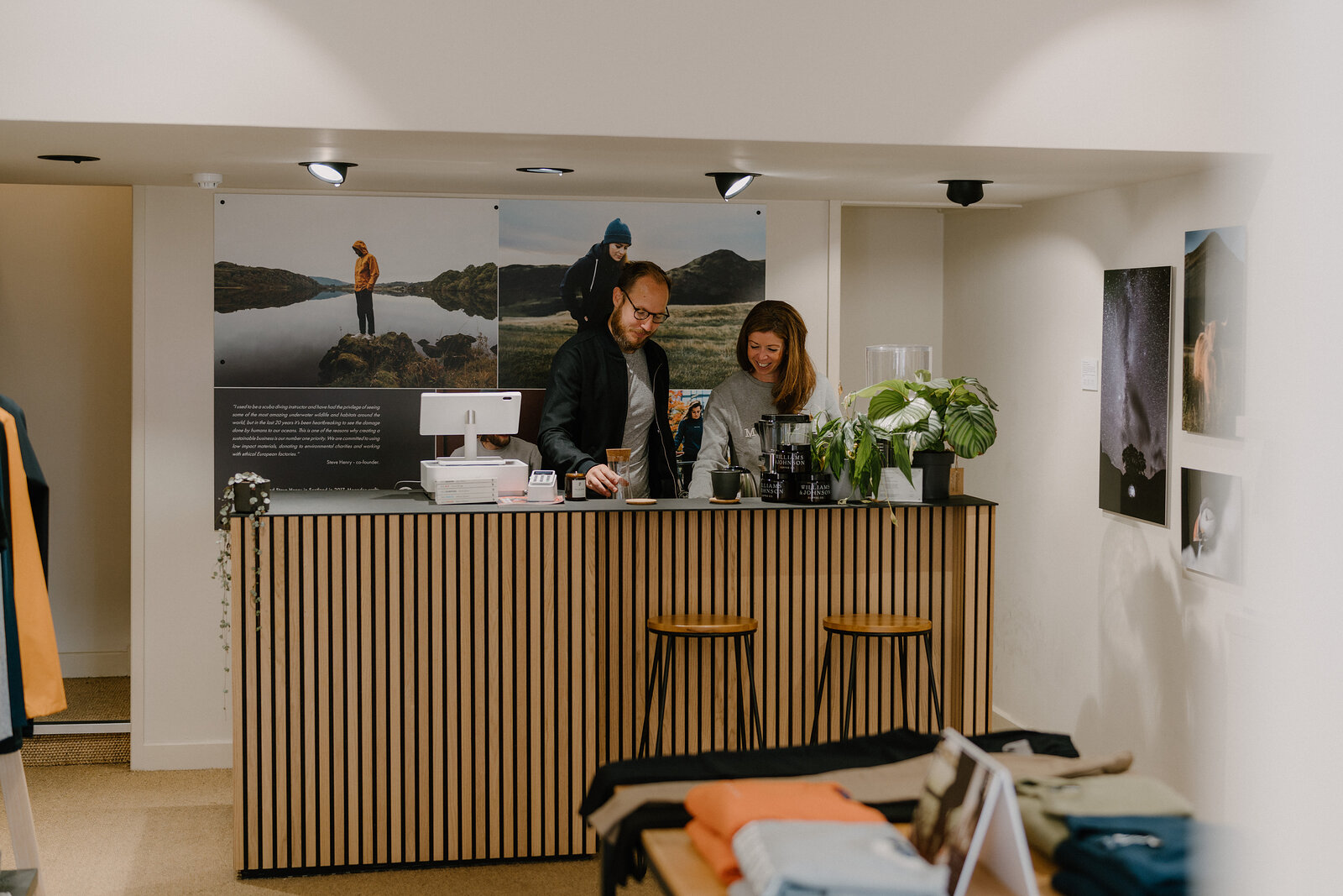 Two people working at a wooden slatted counter beneath a mountain photograph, a laptop and till between them