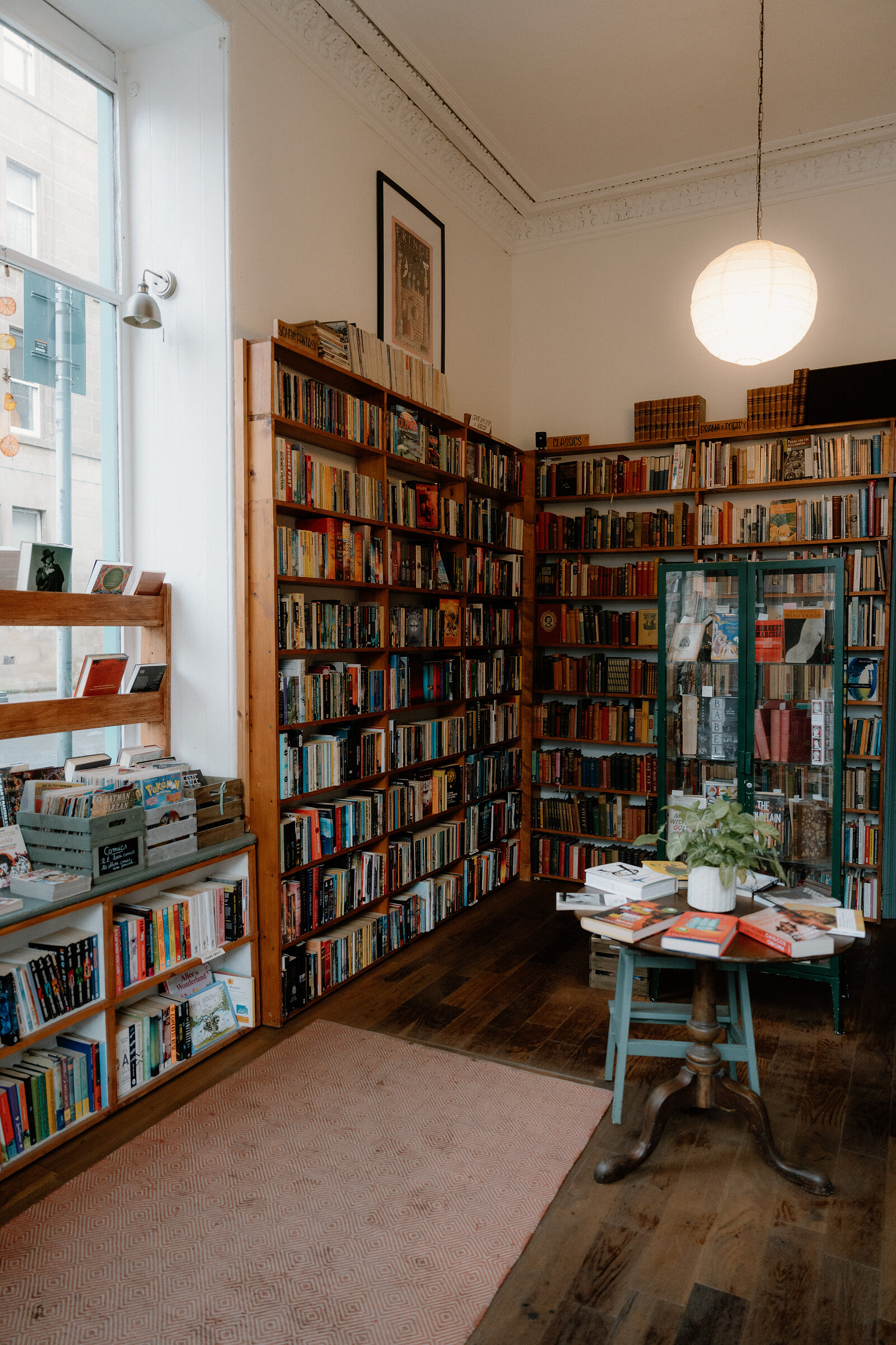 A book-lined room with a glass-fronted cabinet and a round table stacked with titles beneath a paper lantern