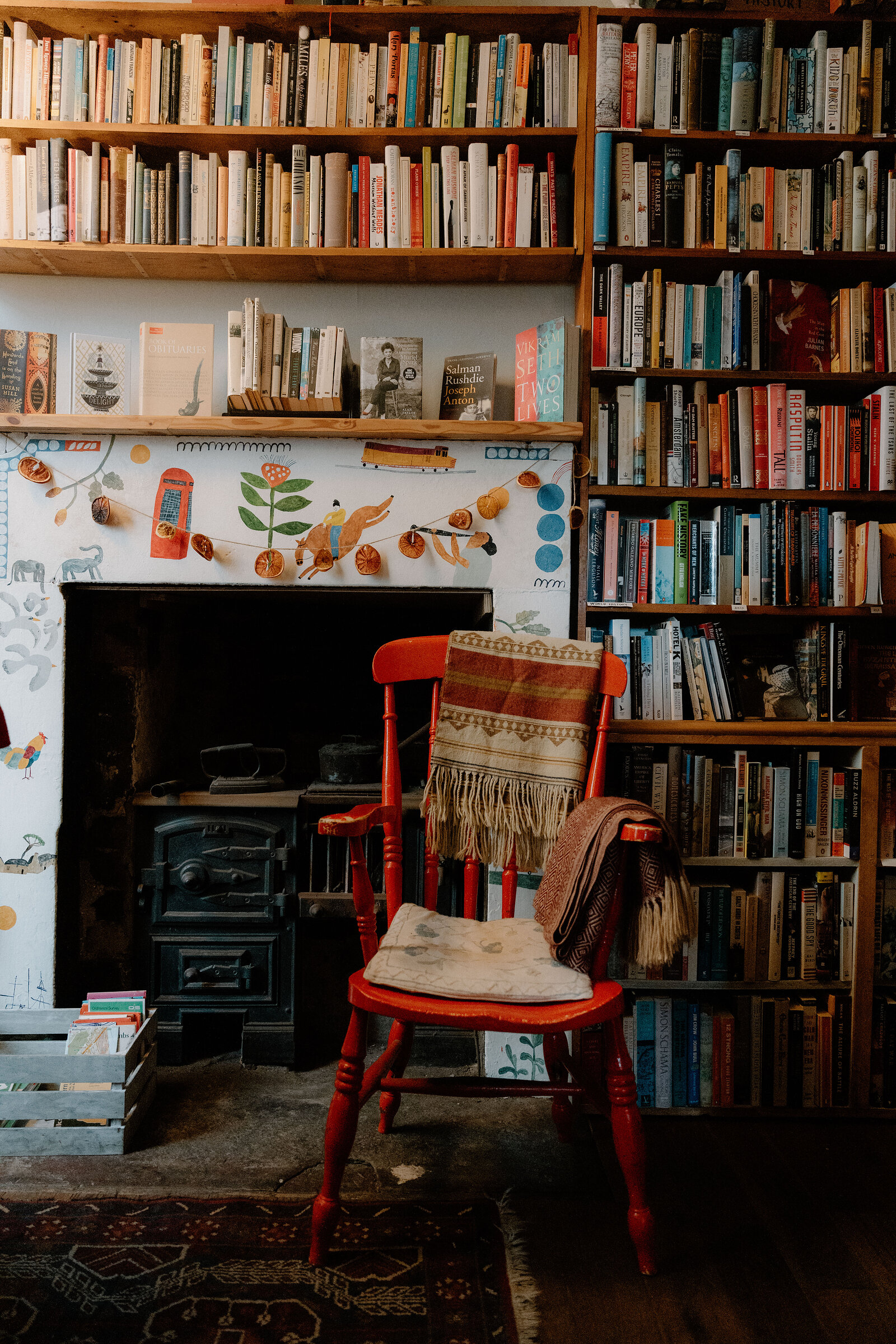 A red wooden chair draped with a throw beside a lit stove, tall bookshelves rising around a painted fireplace surround