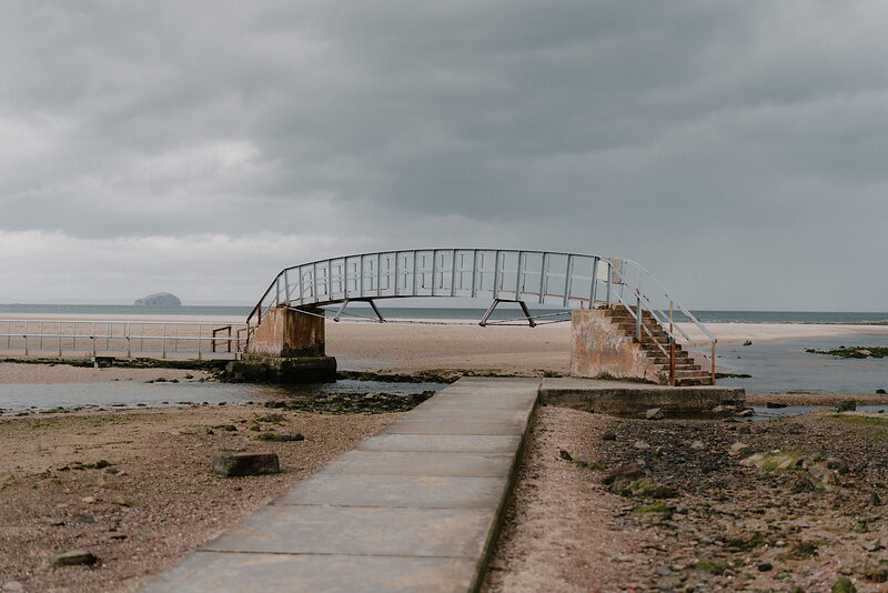 A rusted metal footbridge and steps lead across a sandy beach at low tide under a grey sky, a small stone jetty in the distance.