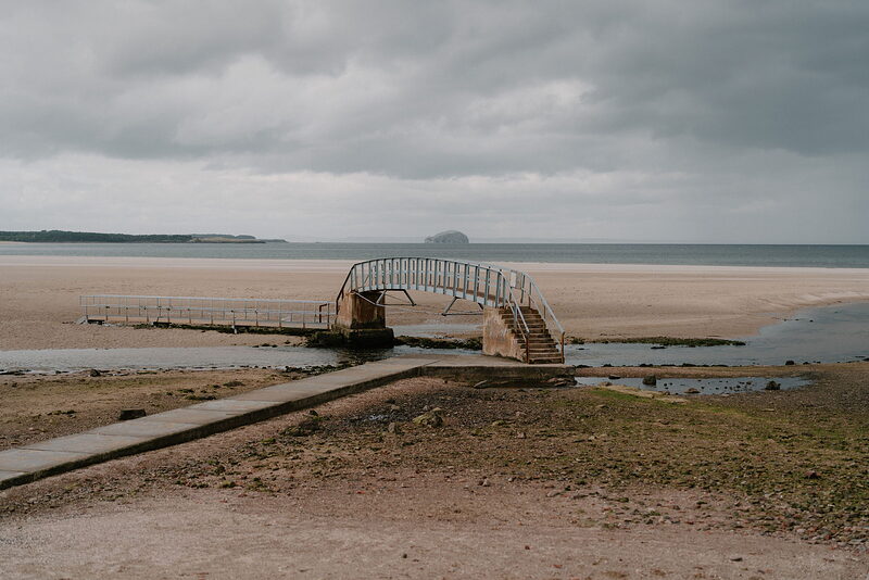 A rusted metal footbridge crosses a shallow channel on a sandy beach at low tide, a distant headland visible under a cloudy sky.