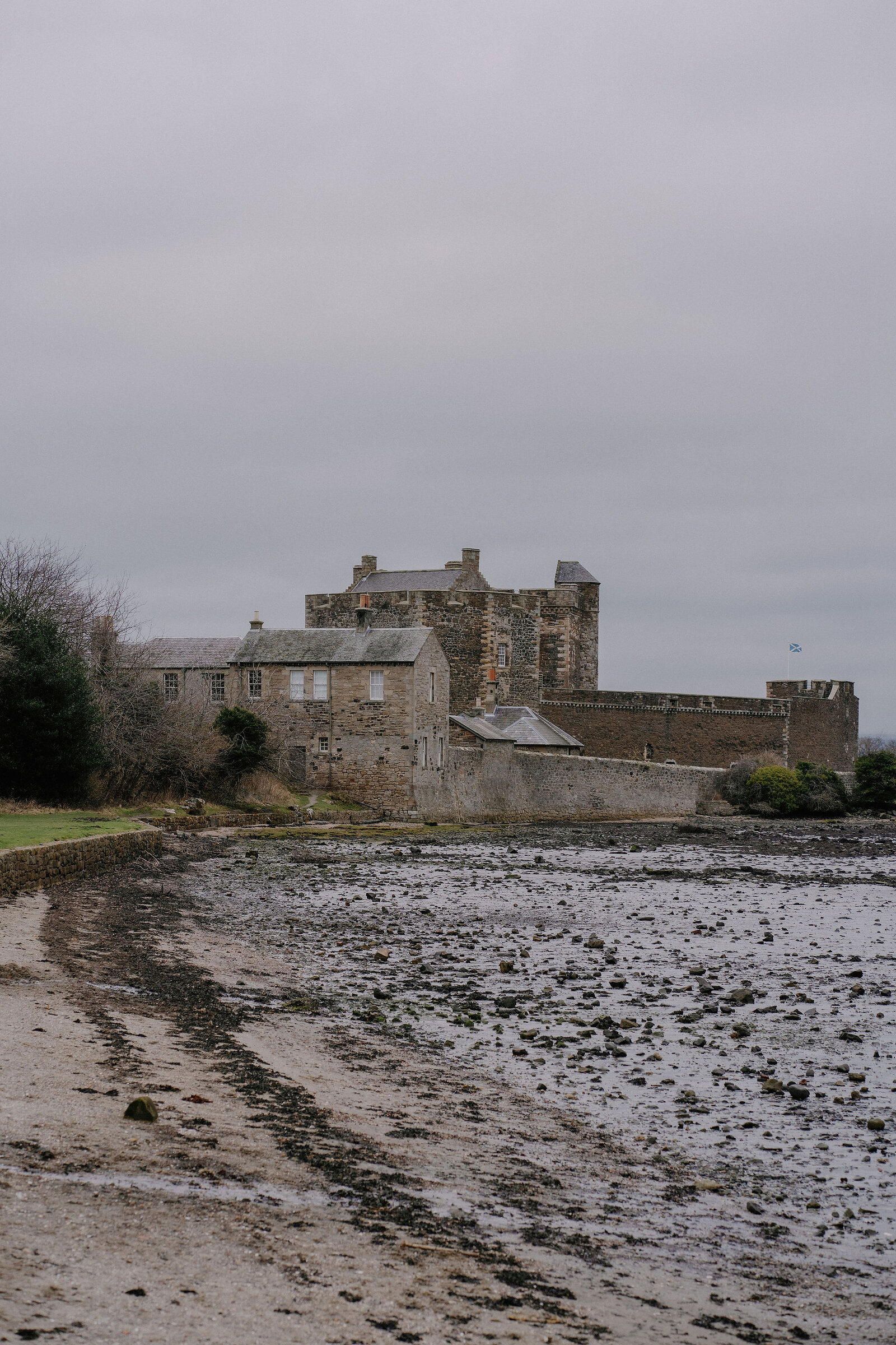 Blackness Castle's keep and battlements rise above a cluster of stone outbuildings beside a muddy shoreline at low tide.