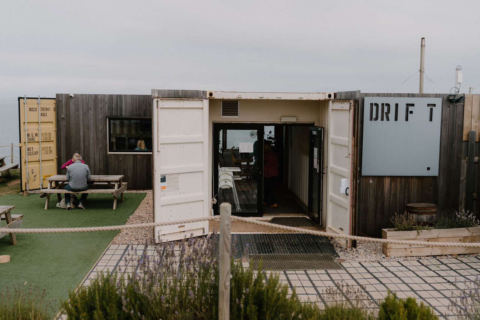 Drift's own sign marks a shipping-container café above the shore, picnic tables outside where people sit looking over the water.