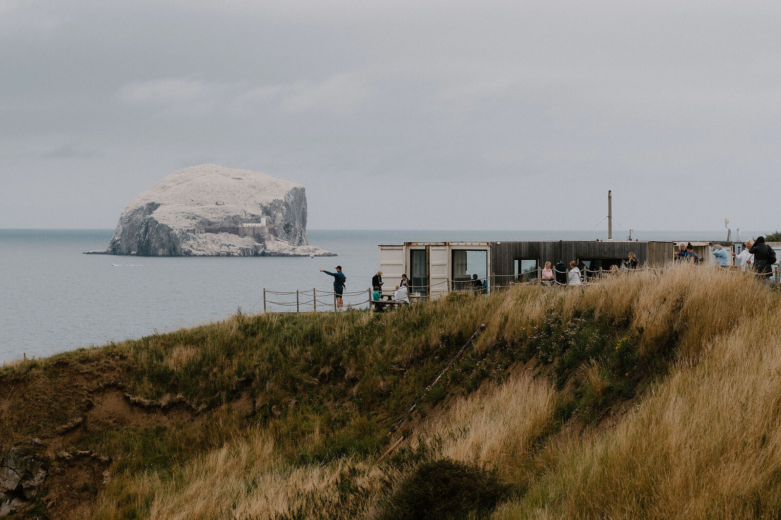 Bass Rock rises from the sea beyond a clifftop visitor building where people gather to look out.