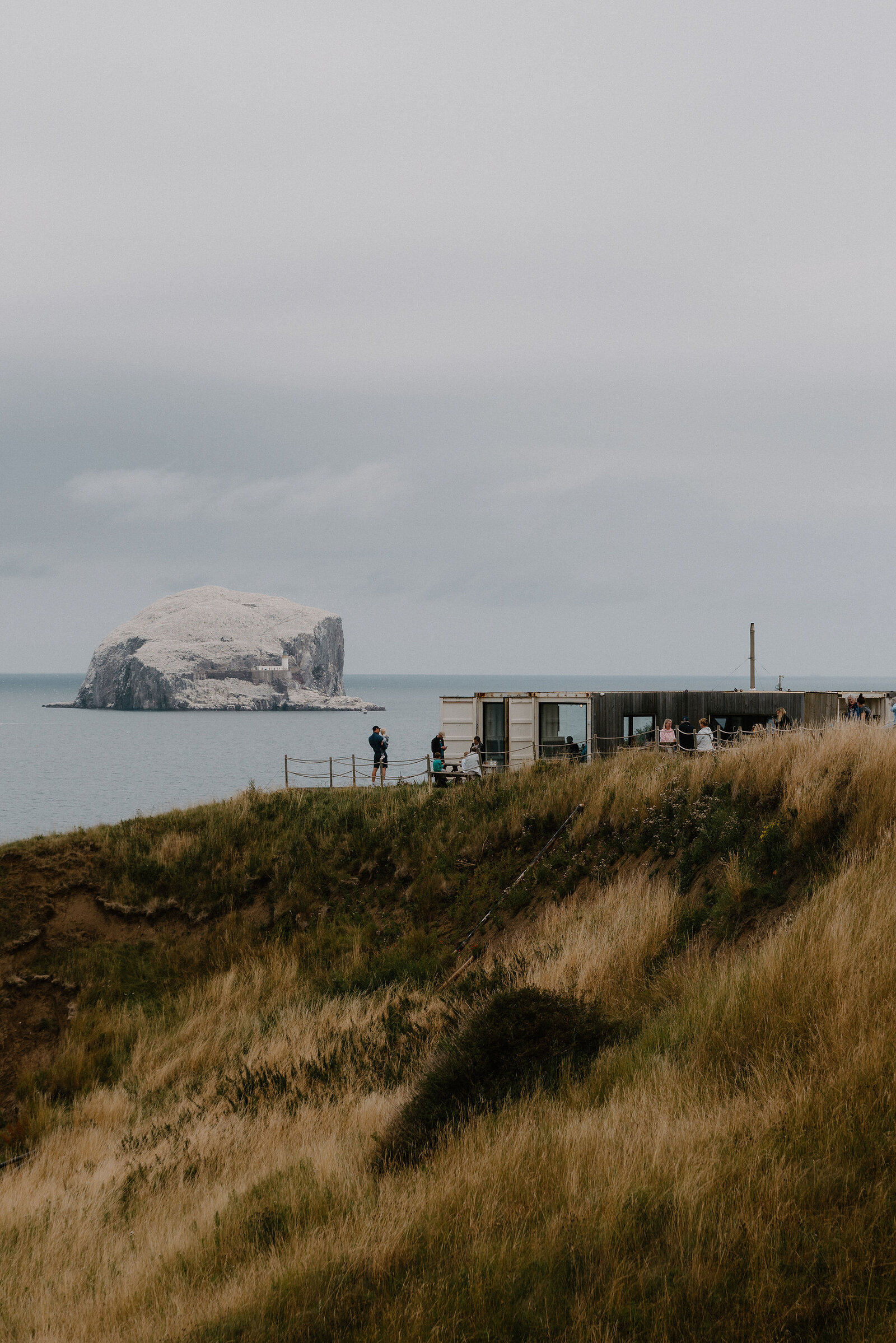 Bass Rock sits offshore beyond a clifftop visitor building where people look out to sea.