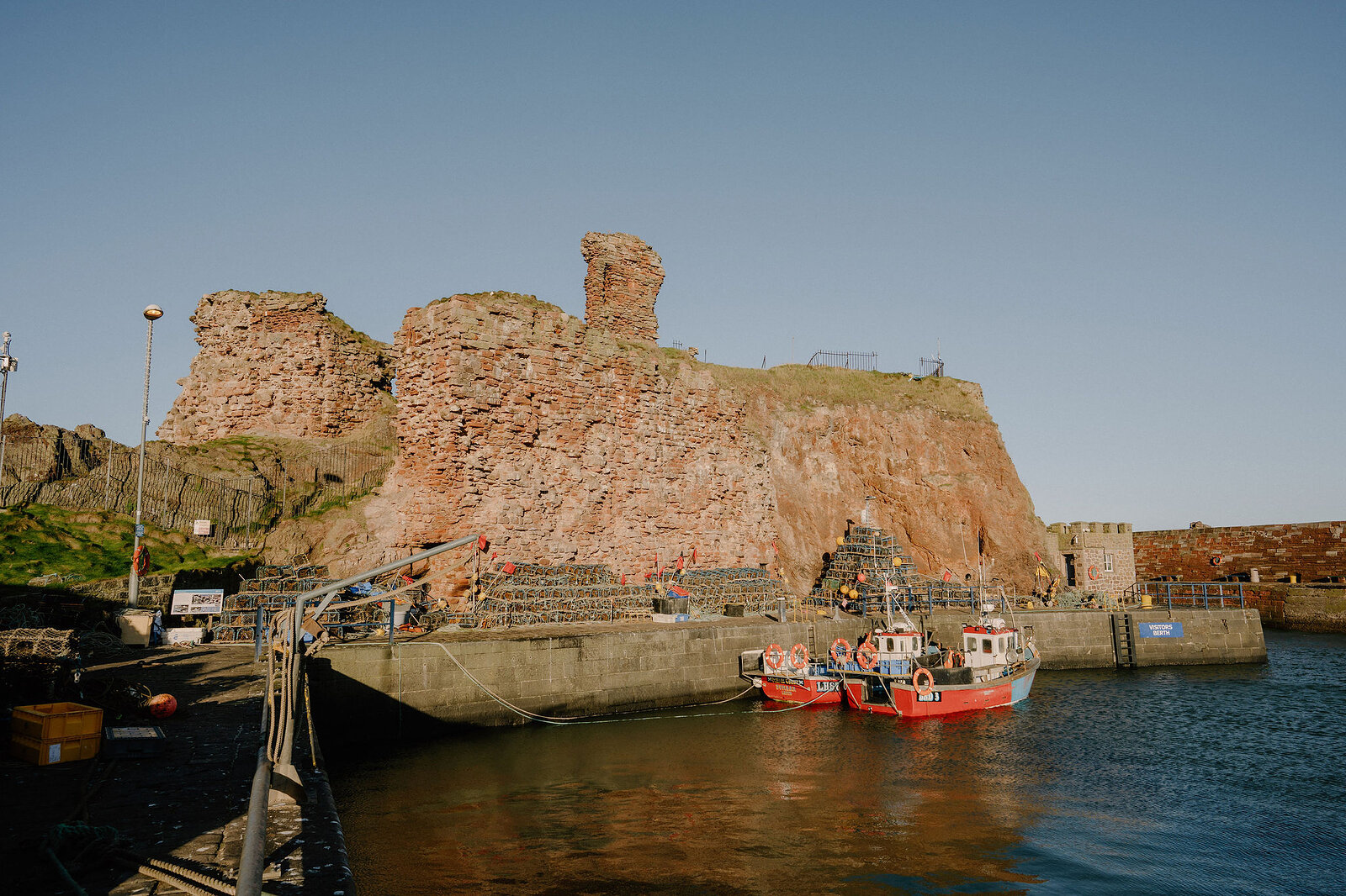 Red sandstone castle ruins rise above a harbour where boats sit moored beside stacked lobster creels.