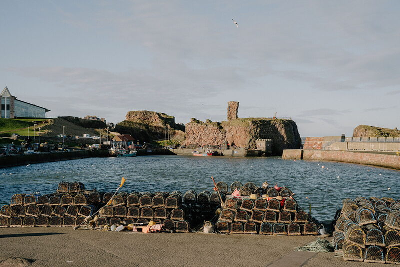 Lobster creels stack on the quay beside boats moored below Dunbar's red sandstone castle ruins.
