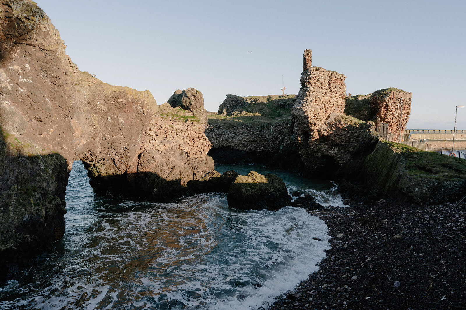 A rocky coastal cove opens beside crumbling stone ruins on the clifftop.