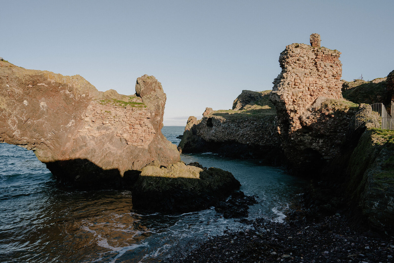 A rocky coastal cove opens beside crumbling stone ruins on the clifftop.