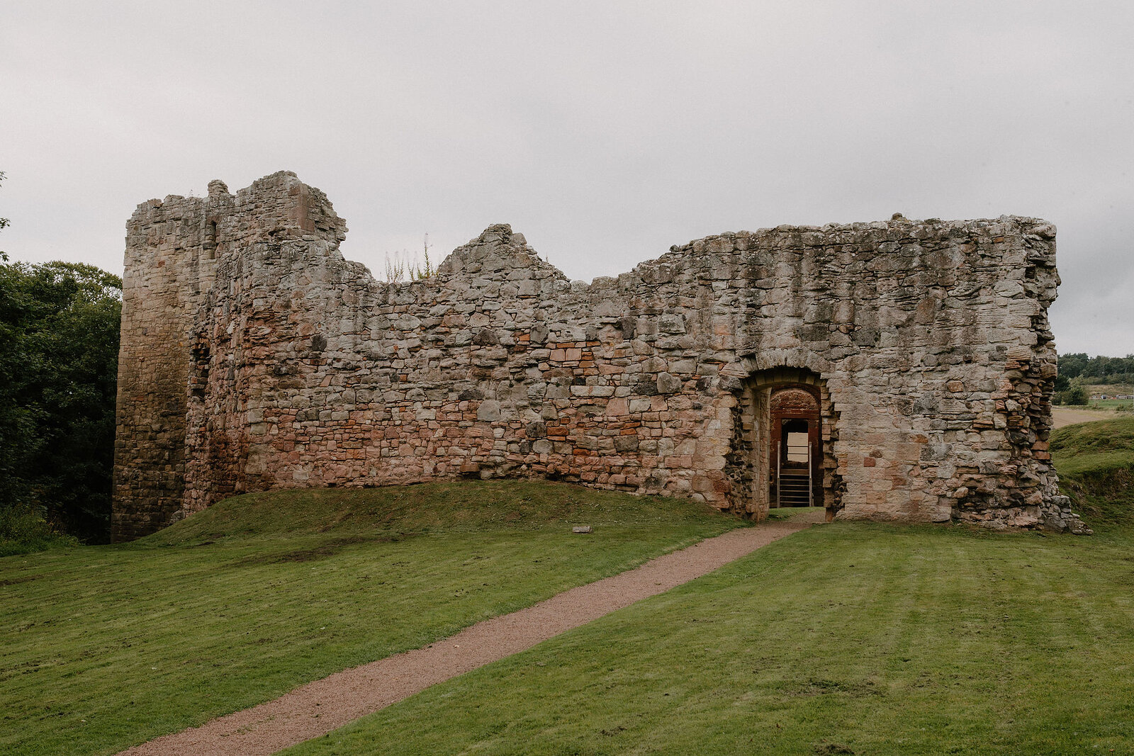 A castle ruin's stone wall stands with an arched doorway leading through to open fields.