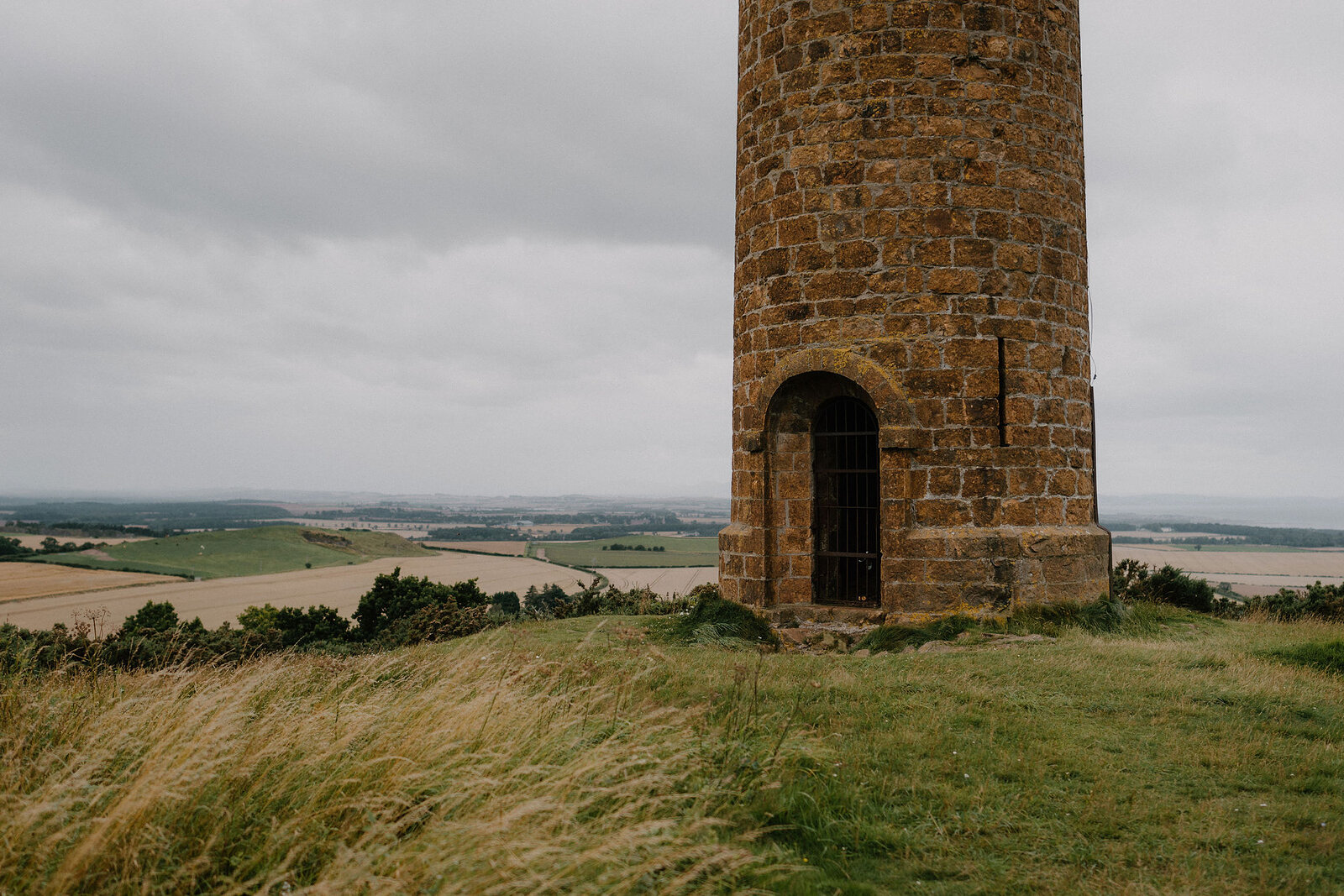 A tall stone tower's arched doorway sits at its base among grass and bushes.