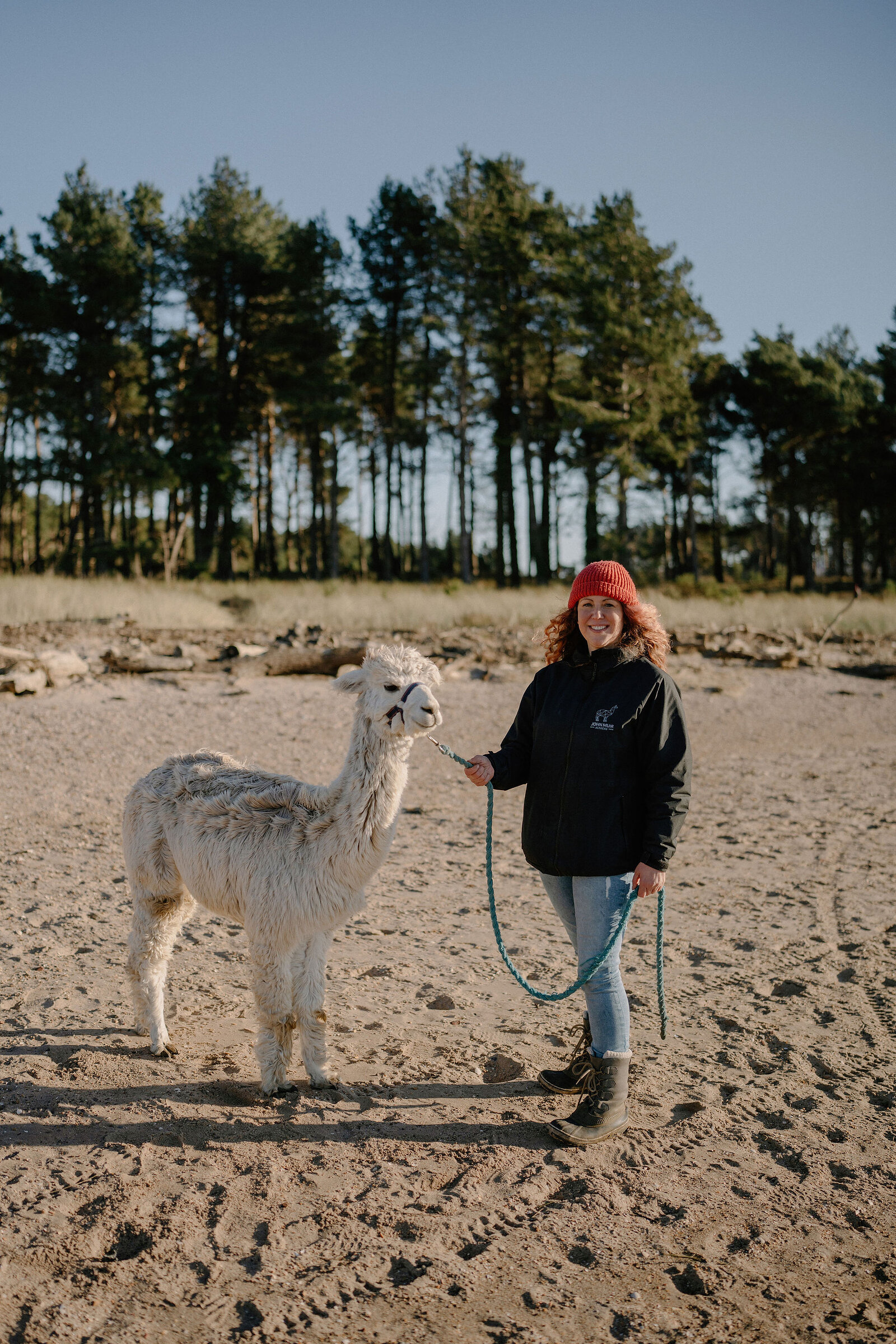A smiling woman in a red hat stands beside an alpaca on a sandy beach.