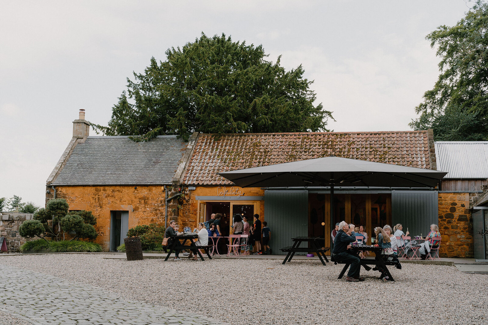 People sit at outdoor tables under a large umbrella beside stone farm buildings.