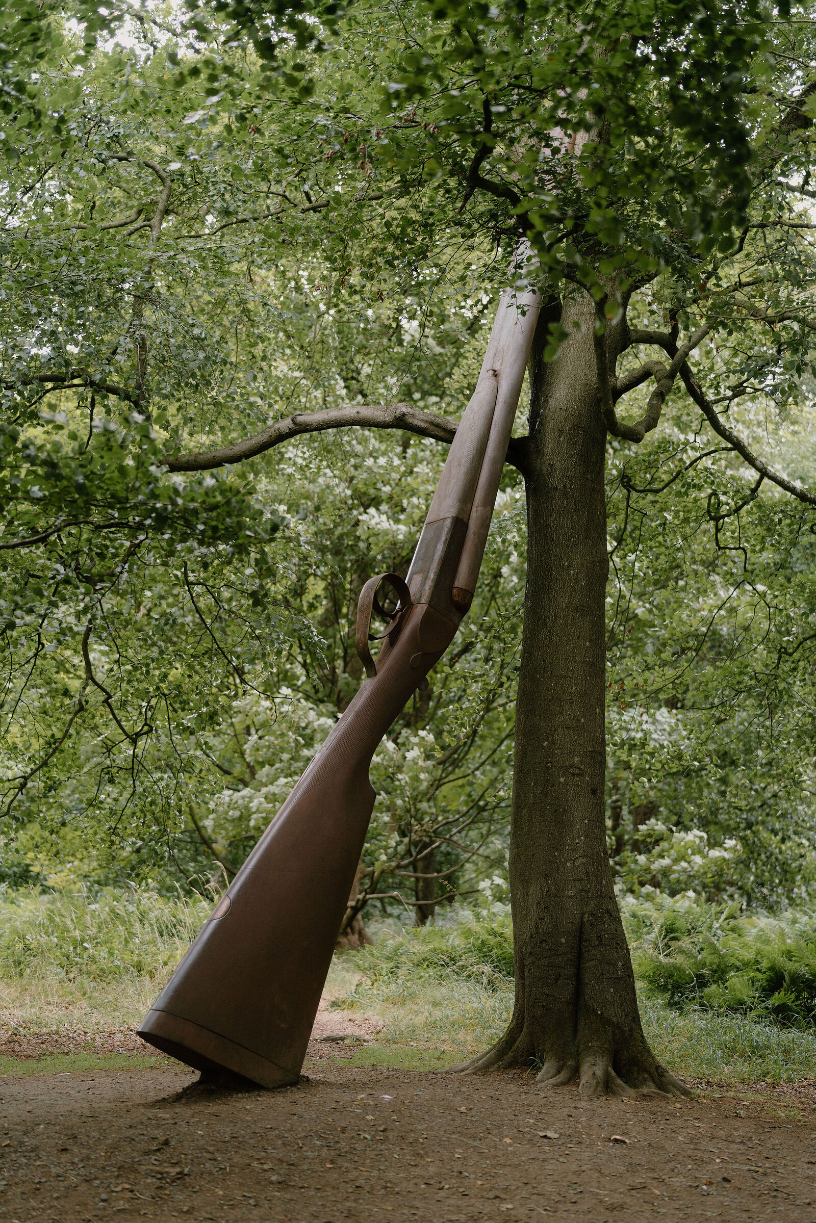 A large sculpture shaped like a rifle leans against a tree trunk in woodland.
