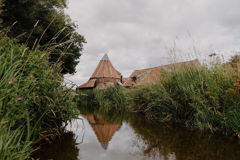 A conical-roofed dovecote reflects in a still stream fringed with reeds.