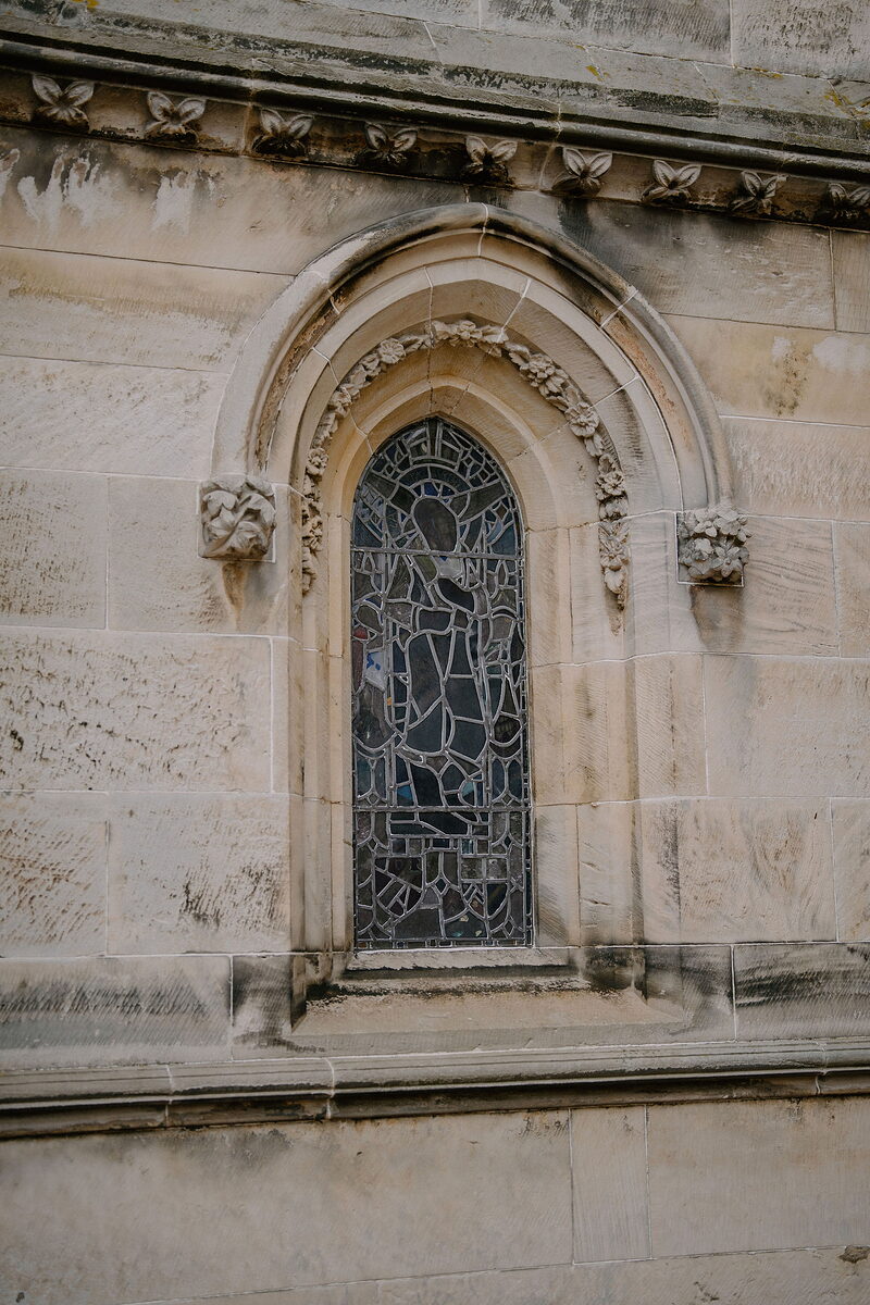 A stained-glass window sits beneath a carved gothic arch on a stone facade.